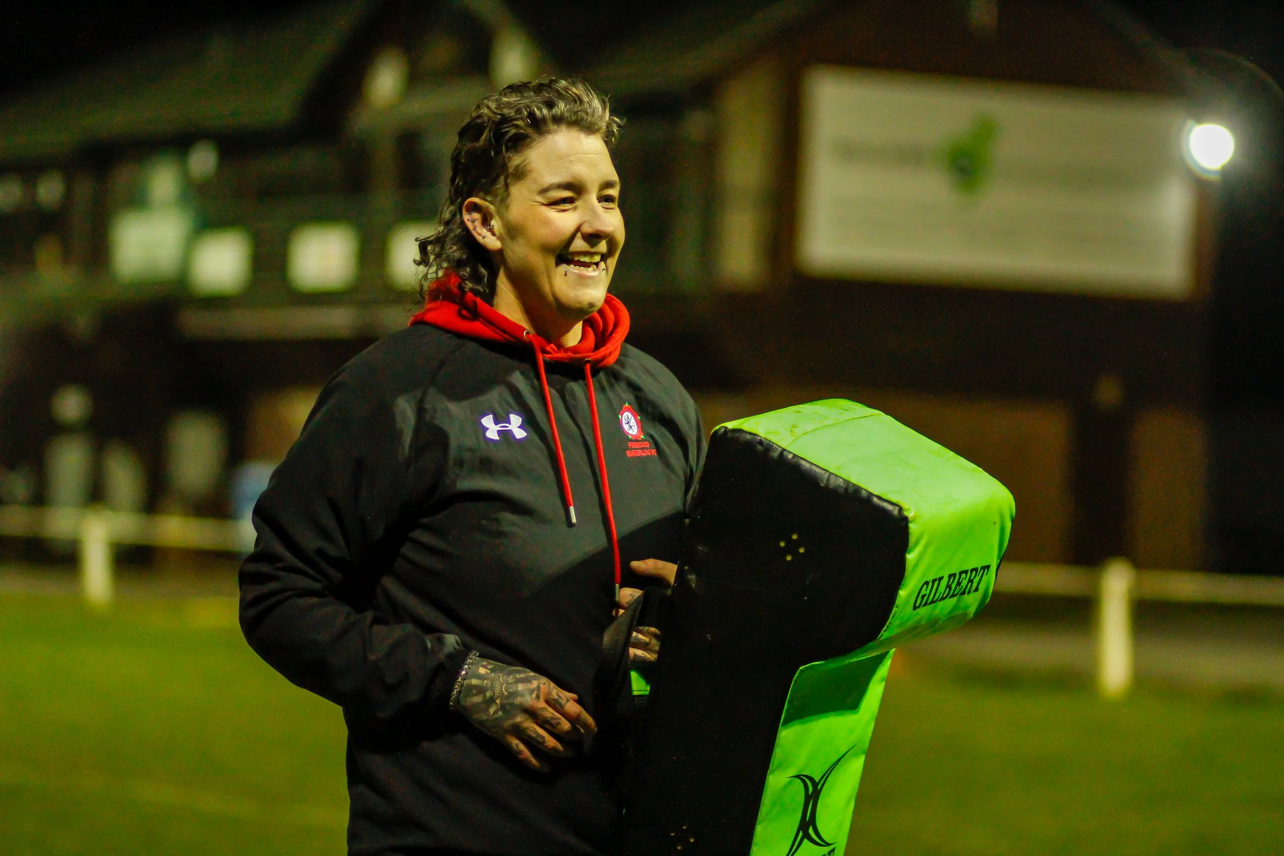 a female coach holding a green and black padded tackling dummy on a rugby field at night, smiling, dressed in black sportswear with red accents.