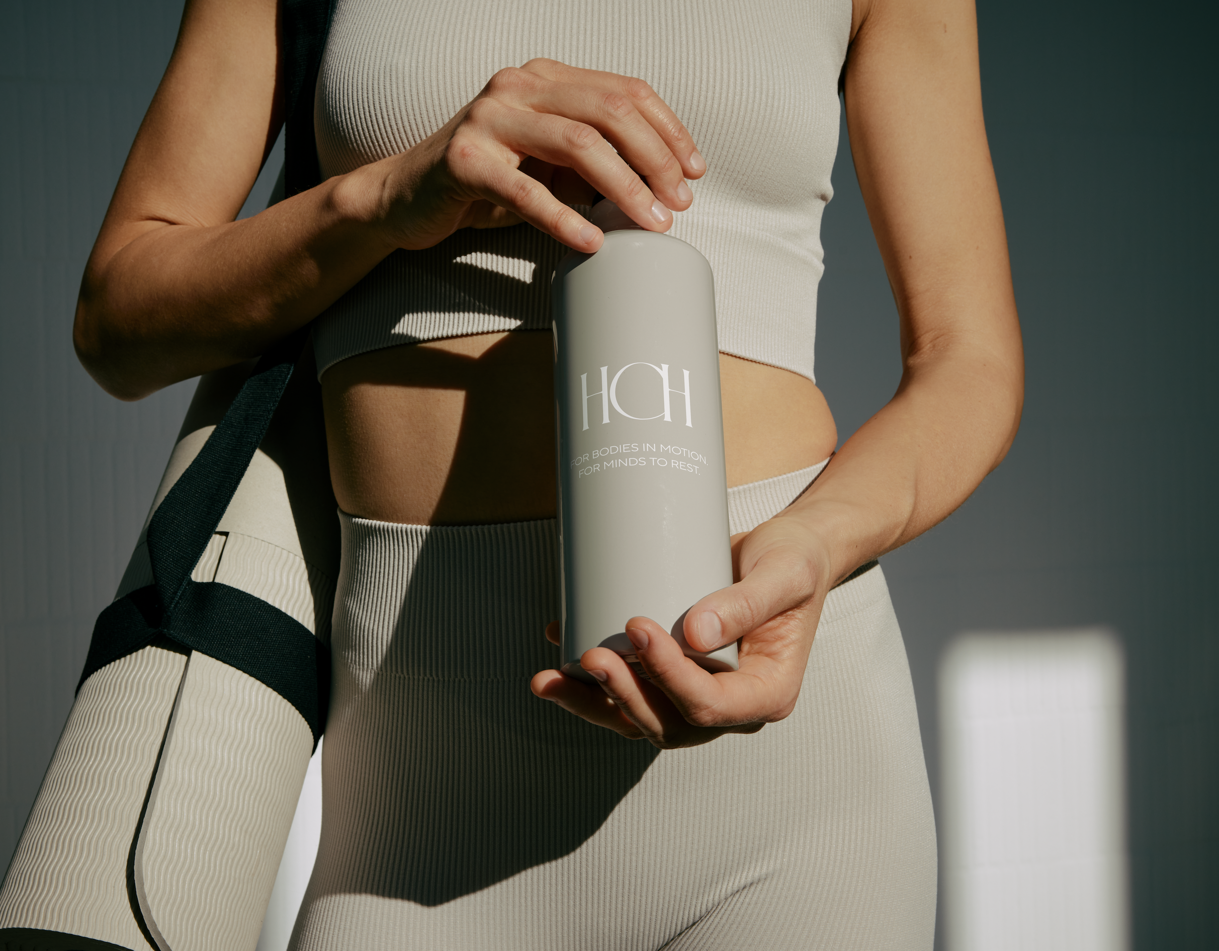 A woman in athletic clothing holding a bottle with the letter 'H' and text that reads 'for bodies in motion, for minds to rest' in front of a plain background.