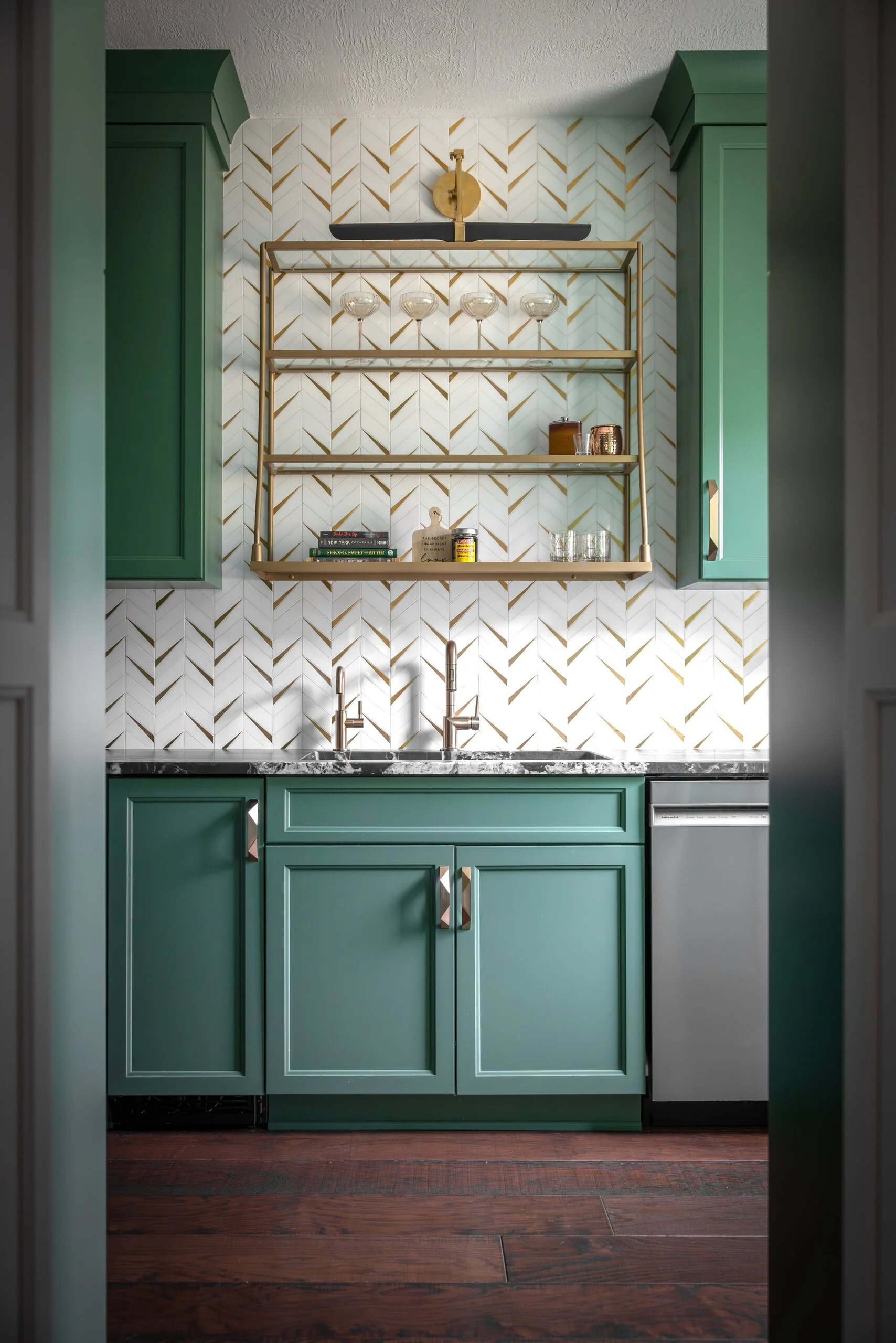Butler's pantry with green cabinetry, gold herringbone tile backsplash, open brass shelving for glassware and a second sink