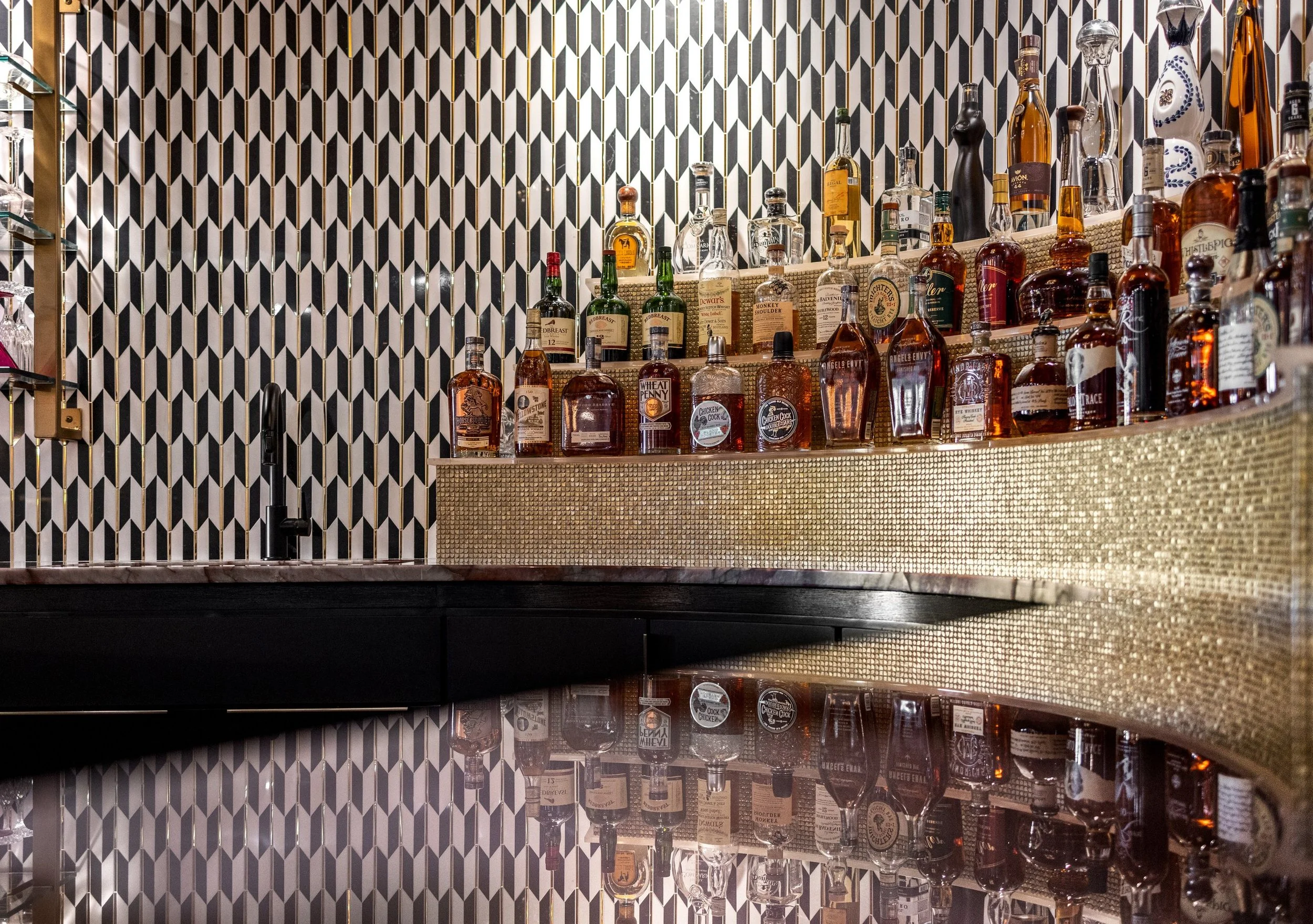 Home bar back shelf wrapped in gold mosaic tile with bottles reflected in a polished counter
