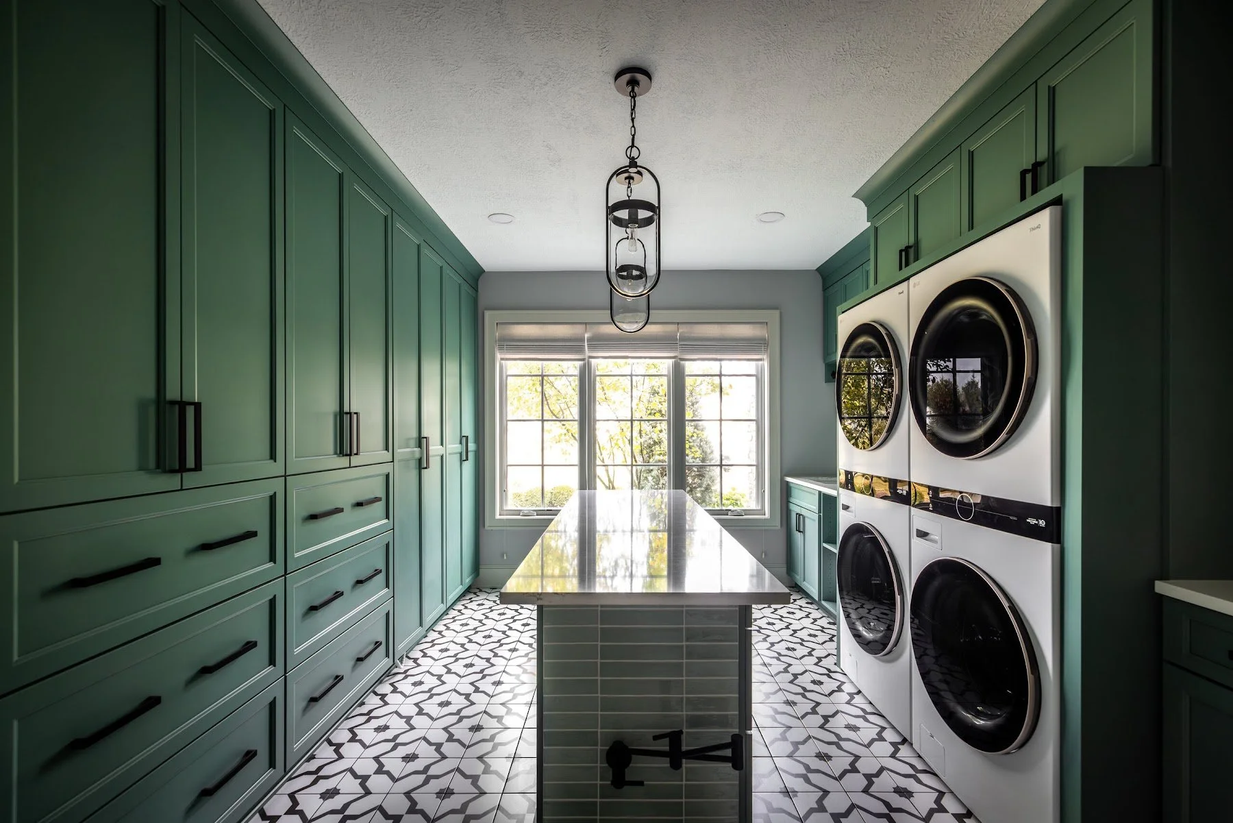 Laundry room with deep green cabinetry, a long folding island, patterned tile floor and stacked washer and dryers