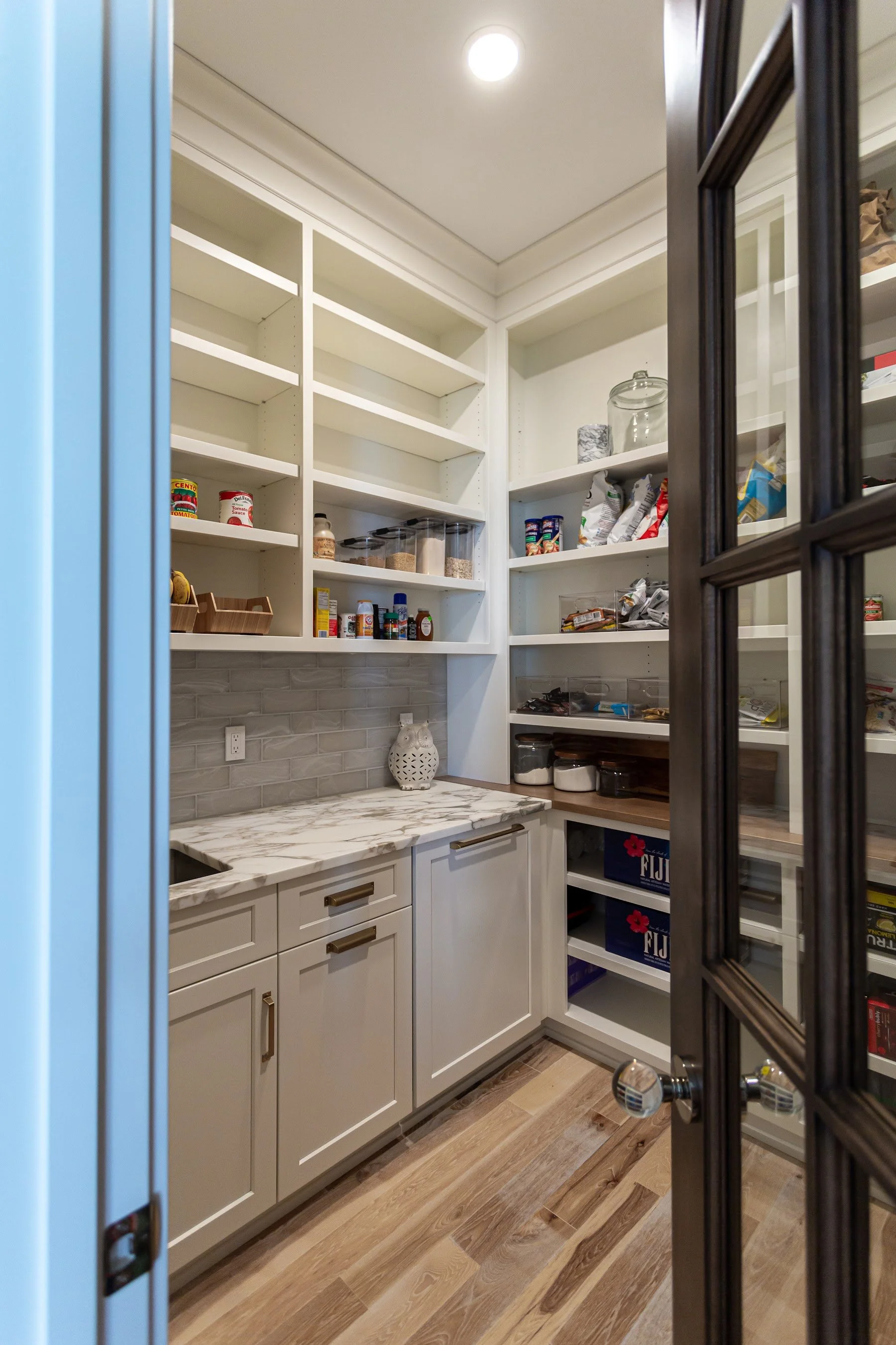 Walk-in pantry behind a black glass-paned door with open shelving, marble counter and lower cabinets for holiday serving pieces