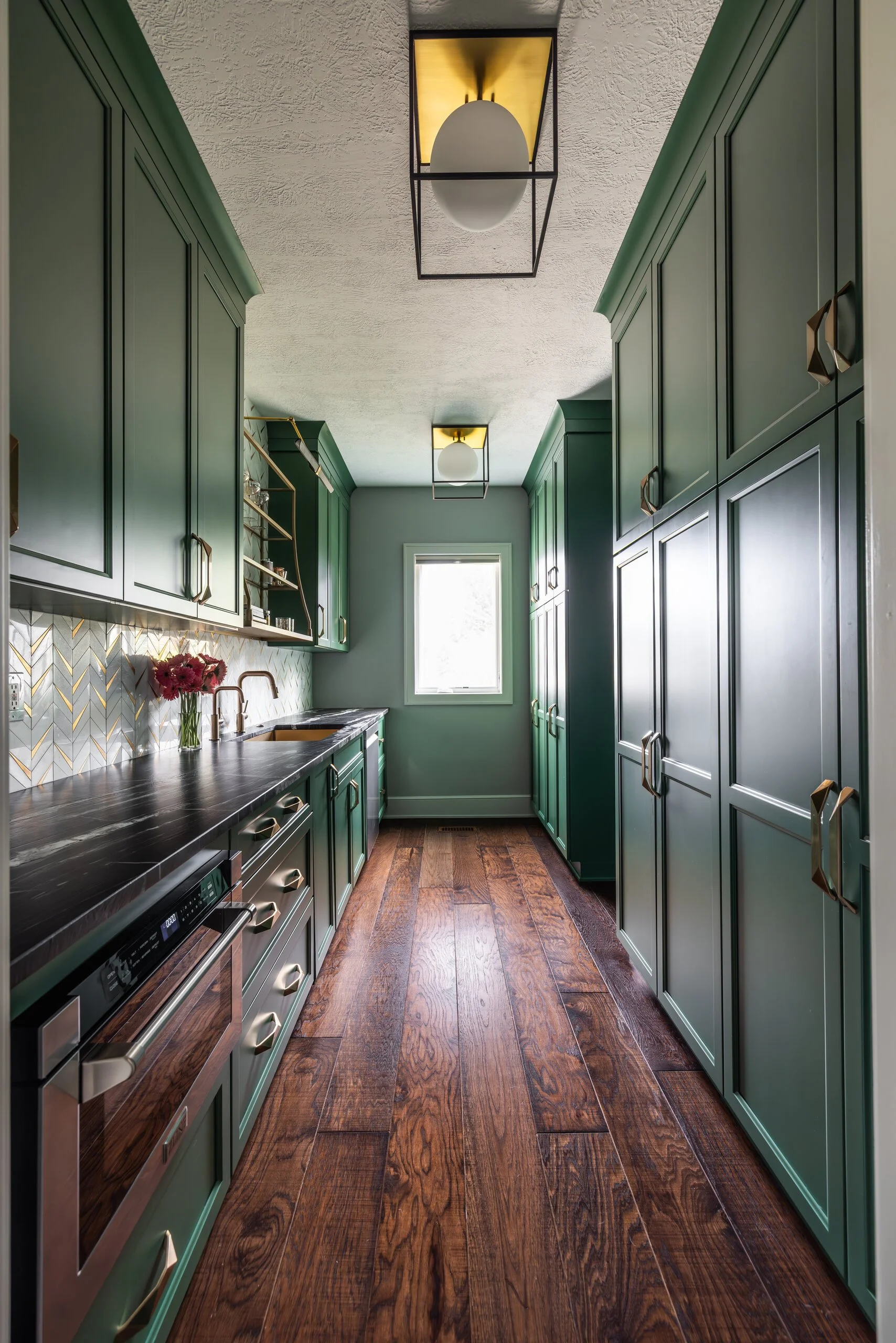Galley scullery with deep green cabinetry, brass lantern pendants and wide plank wood floors behind the main kitchen
