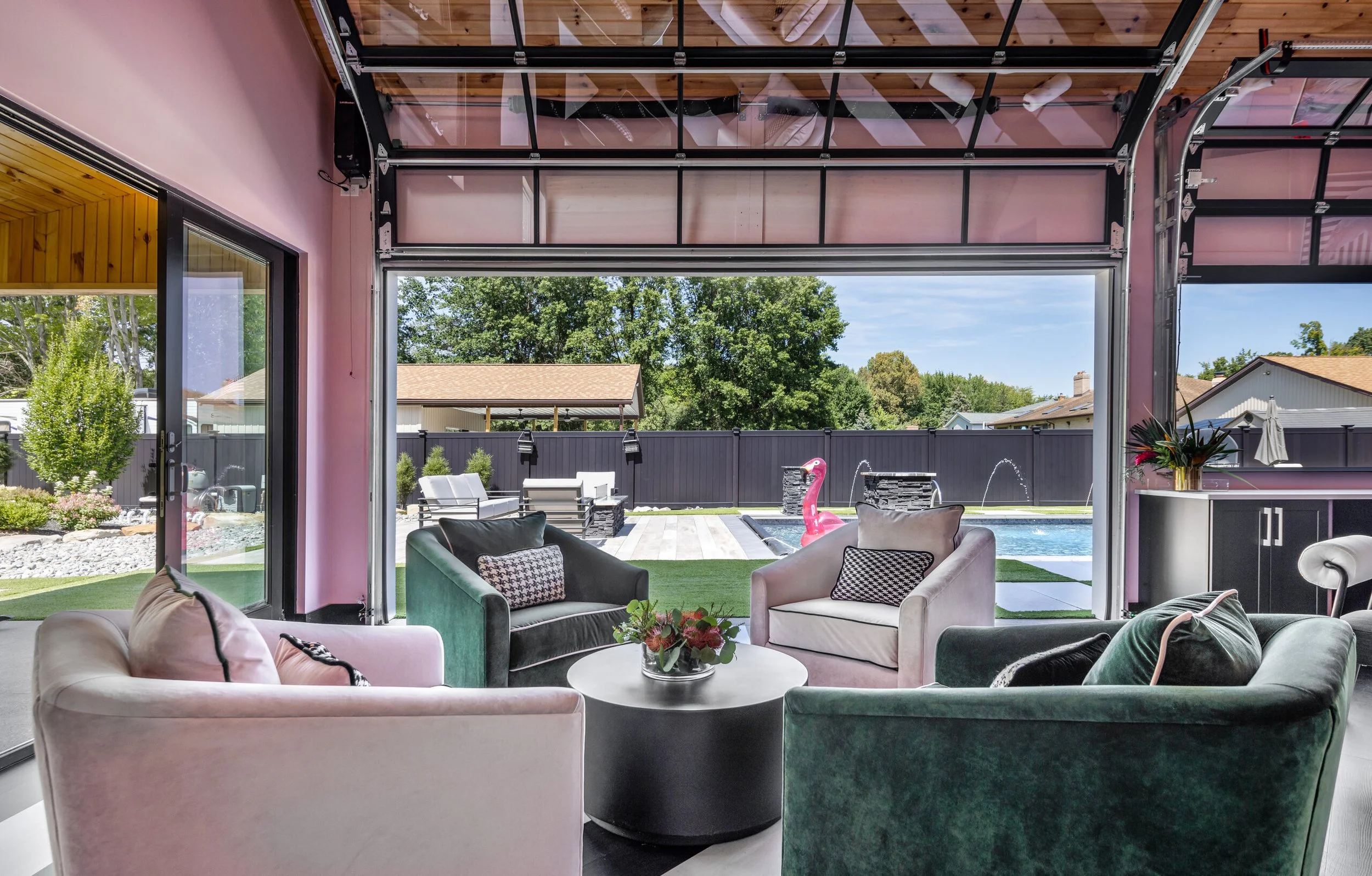 Covered pool house lounge with green velvet sofas, blush pink walls and glass doors open to the pool