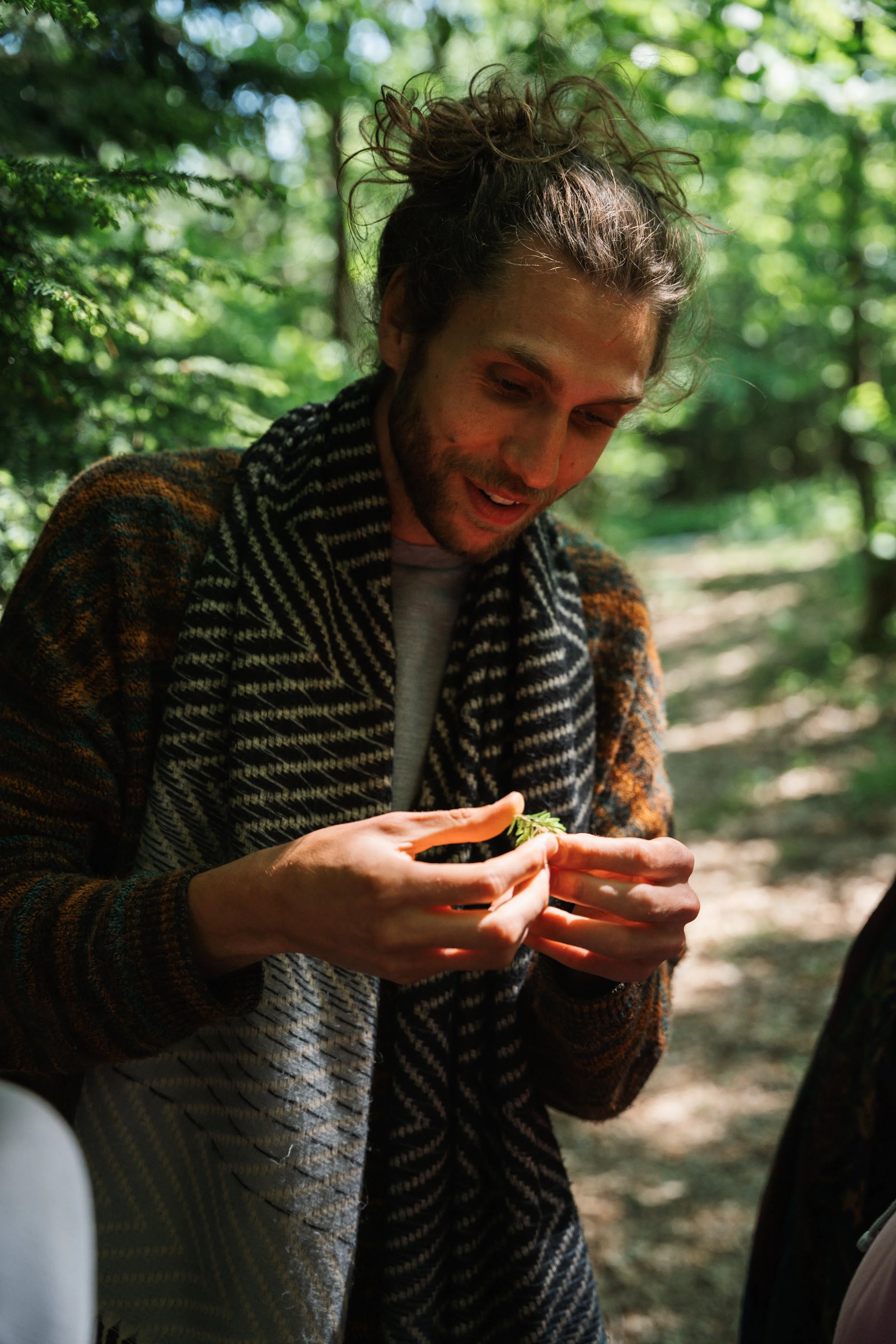 A man with long, curly hair and a beard, wearing a patterned scarf and jacket, is outdoors in a forest, holding and examining a small plant.