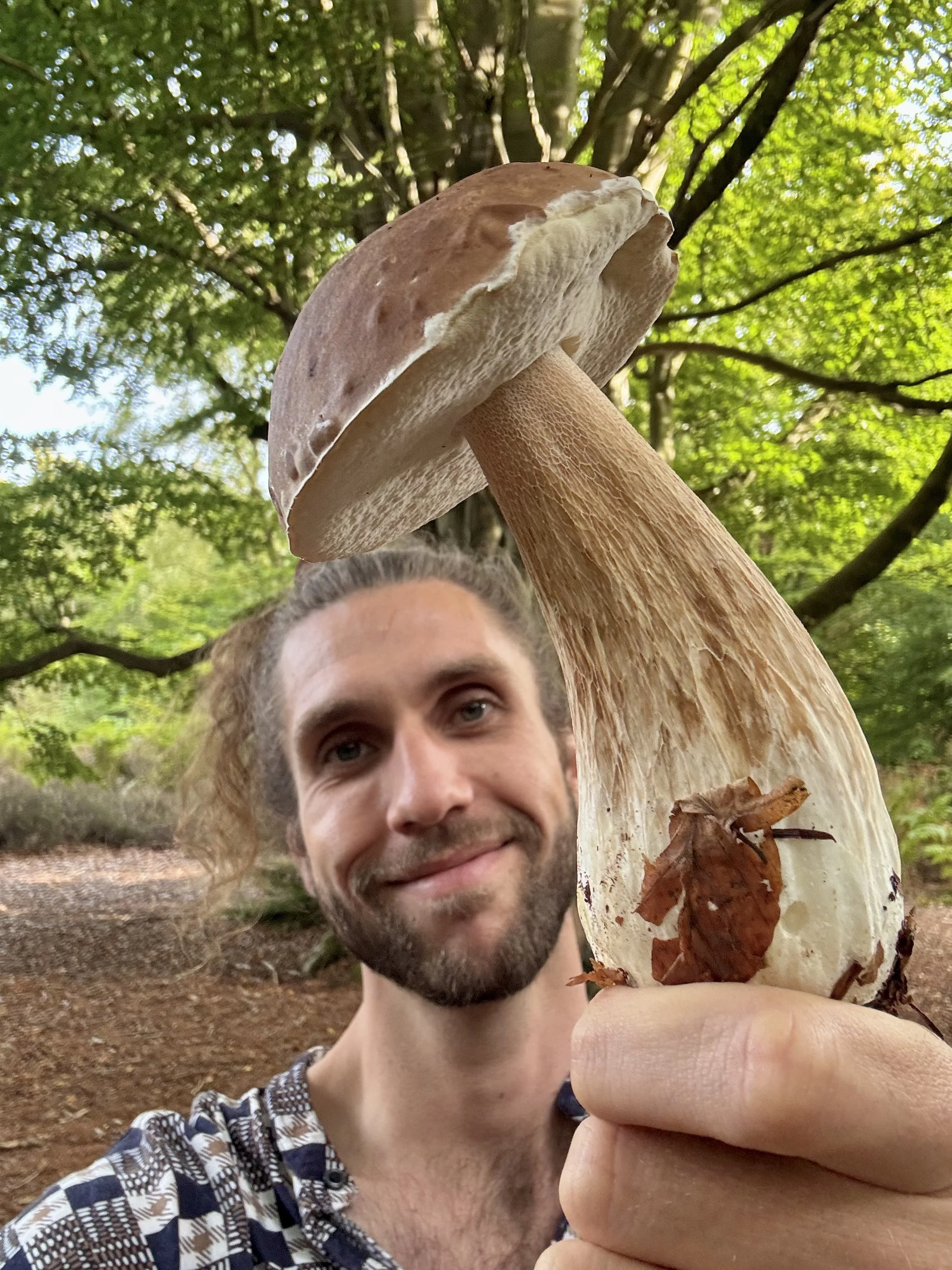 A man outdoors under green trees, holding a large mushroom close to the camera, with a cheerful smile.