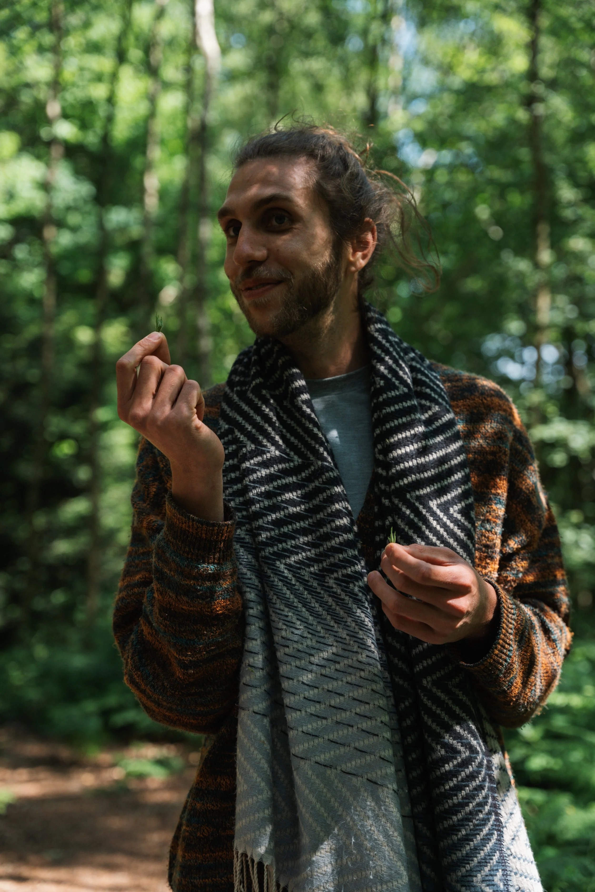 A man with long, curly hair and a beard standing in a forest, holding a small leaf and a piece of plant, wearing a patterned scarf and a colorful sweater.