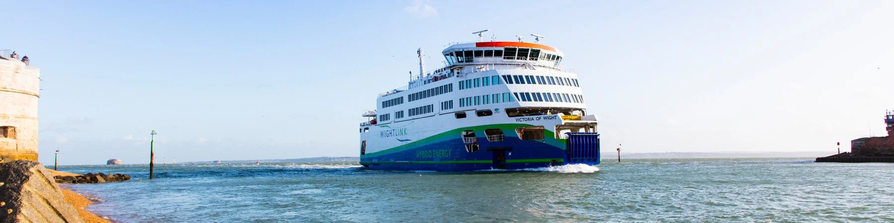 Large passenger ferry boat sailing in a harbor, with a portion of a stone pier on the left and a small lighthouse on the right.