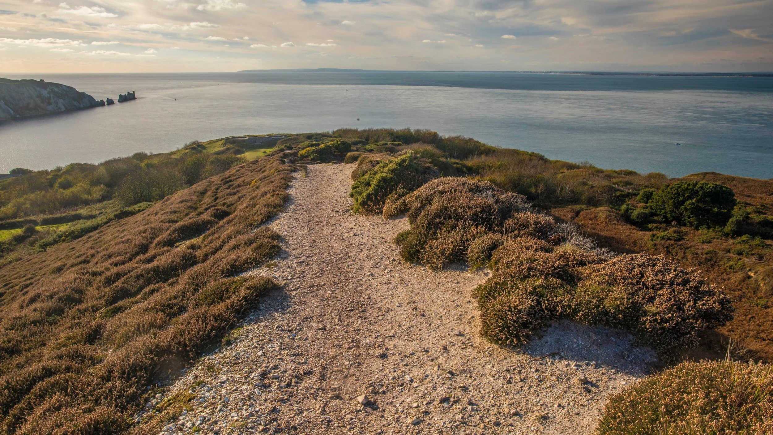 A gravel dirt path along a coastal hillside with shrubbery and bushes, overlooking the ocean under a partly cloudy sky.