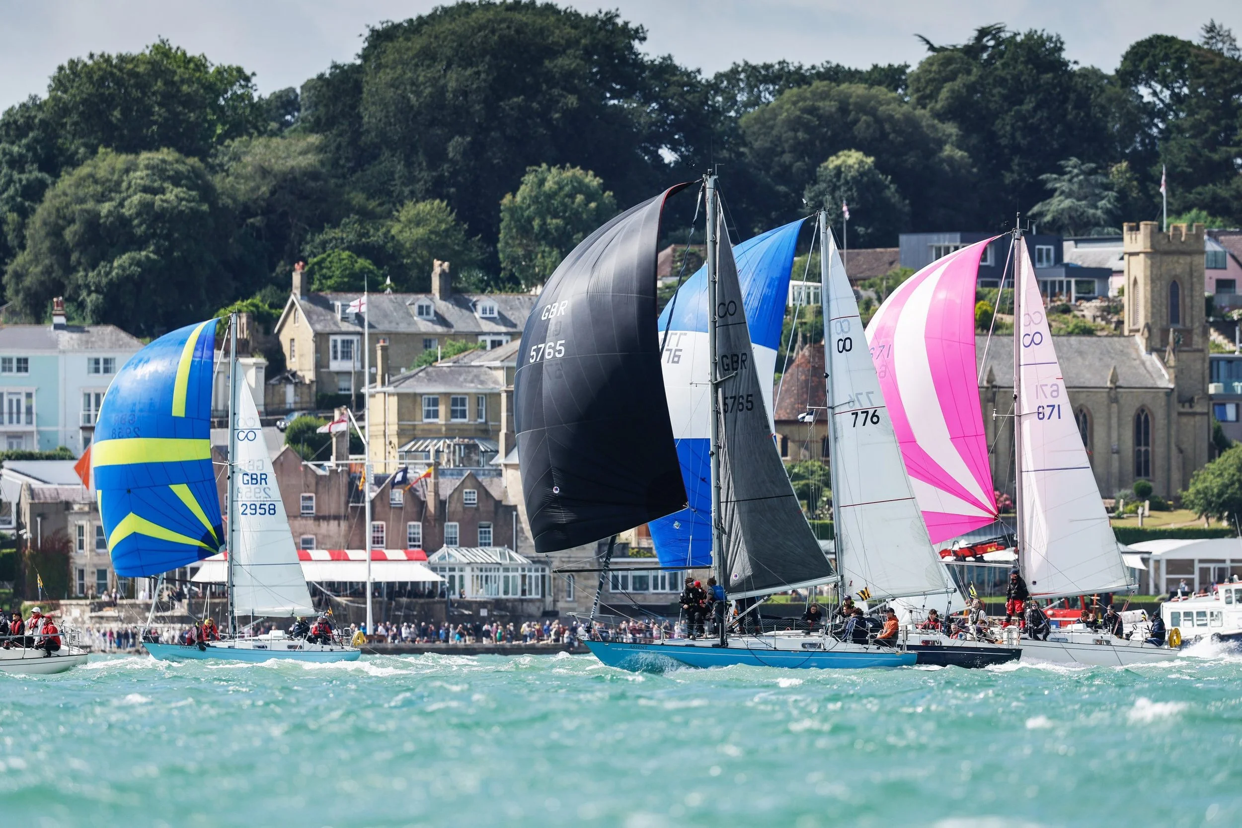 Multiple sailboats with colorful sails participating in a regatta on a body of water, with a waterfront landscape featuring houses and a church in the background.