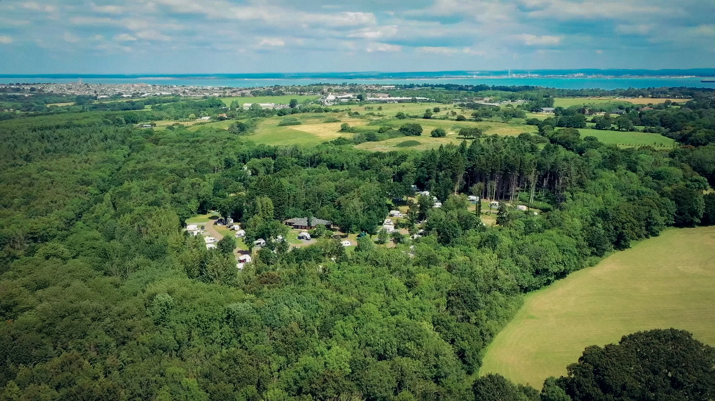 Aerial view of lush green forests, open fields, and a small campground with caravans and tents, with a body of water in the distance under a partly cloudy sky.