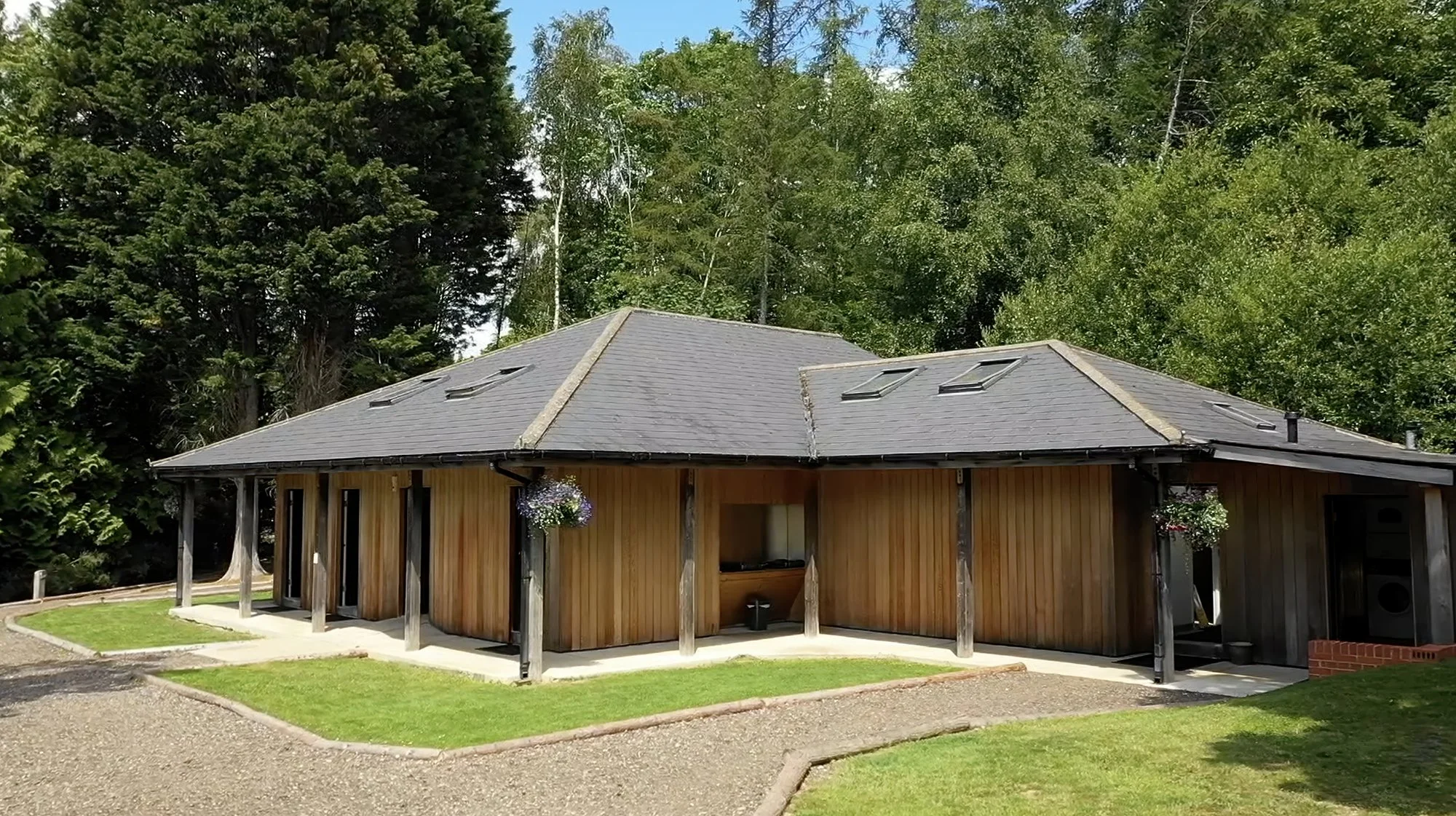 A house with a wooden exterior and a dark gray roof surrounded by greenery, including trees and grass, with hanging flower baskets near the entrance.