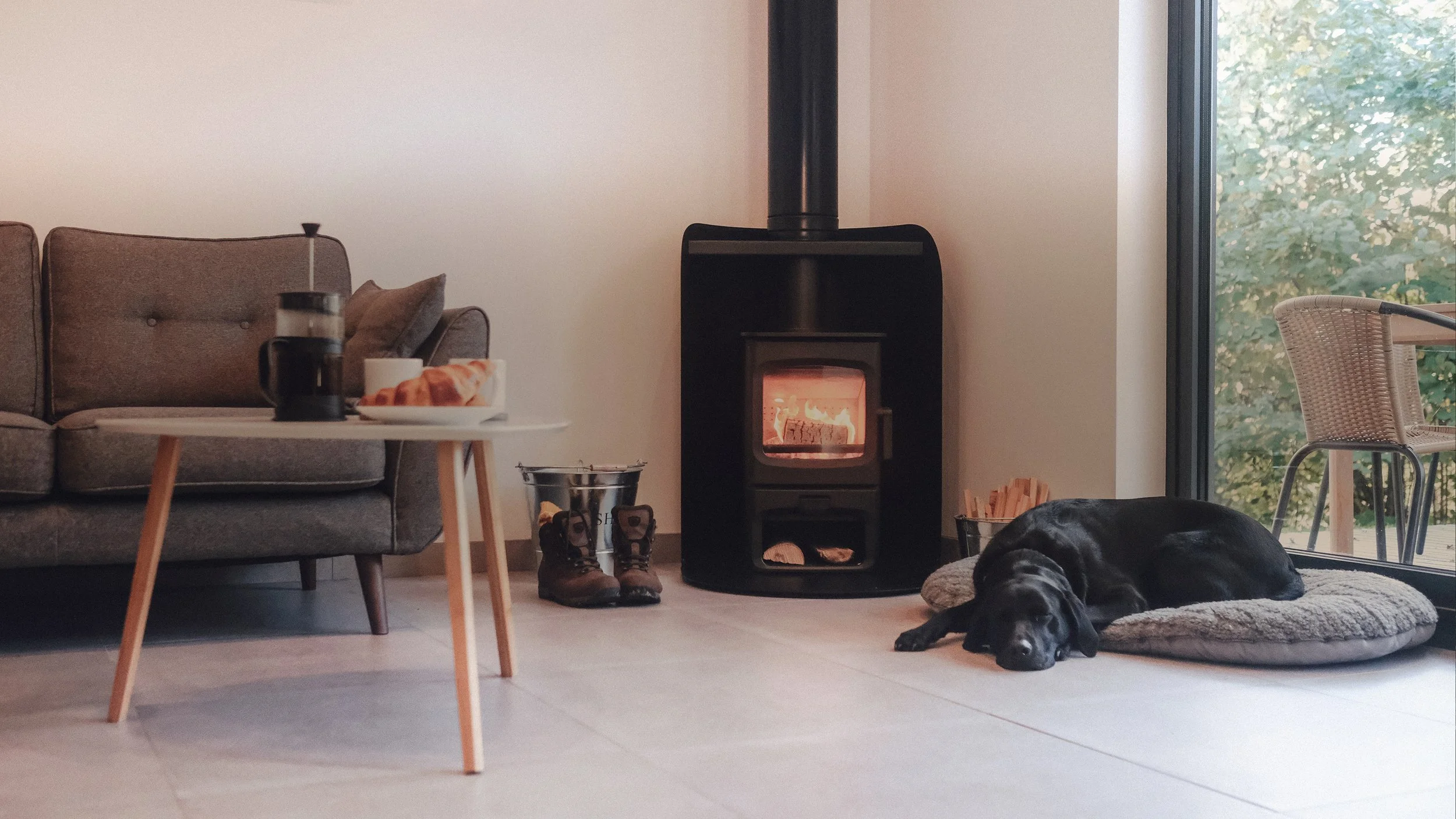 Living room with a black dog lying on a gray cushion near a glass door, a wood stove with visible flames, a small table with a coffee maker and croissants, and a pair of hiking boots on the floor.