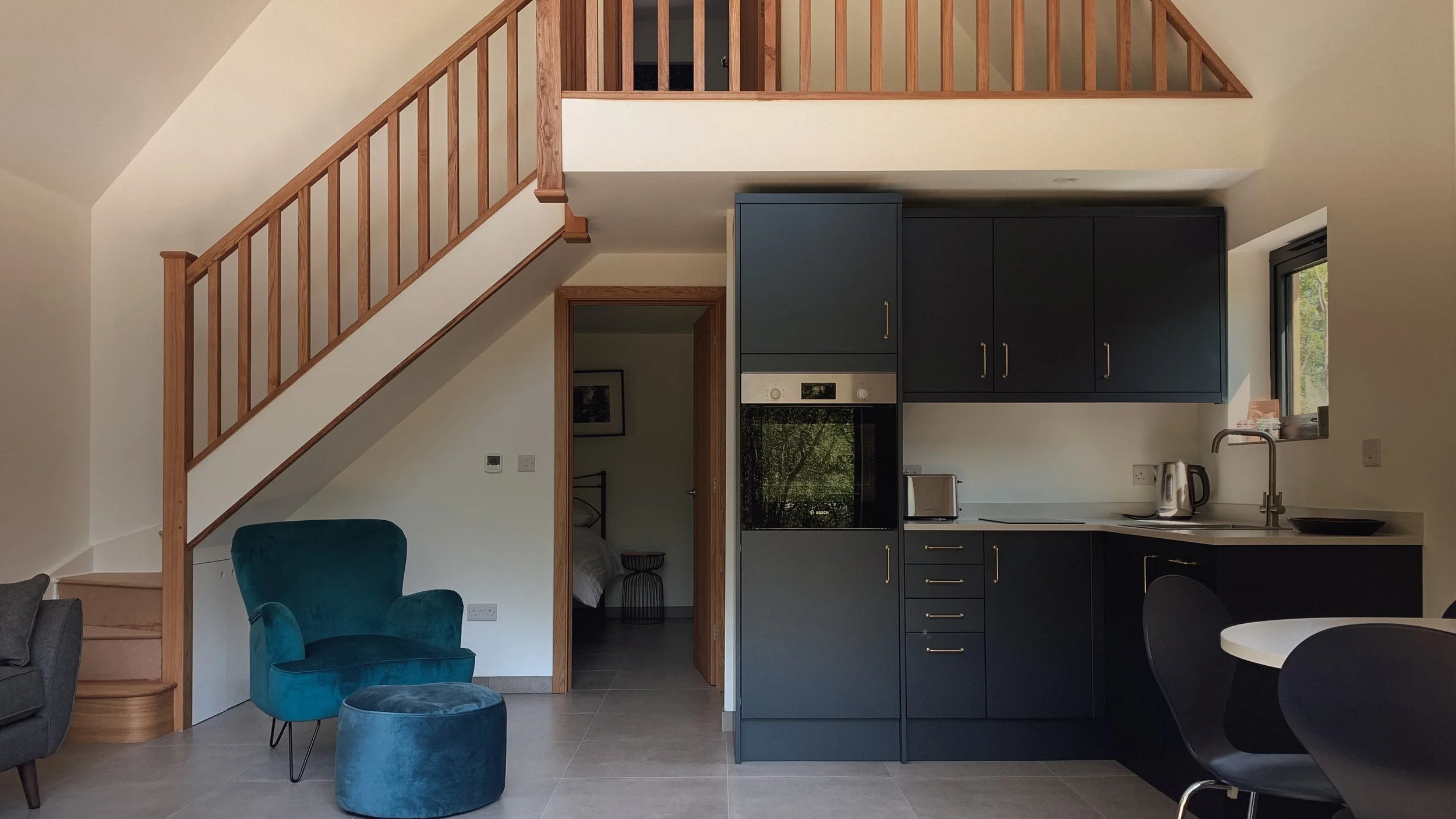 Interior view of a modern living space with a staircase, armchair, kitchen with dark cabinets, oven, toaster, kettle, and a window, with partial view of a bedroom.