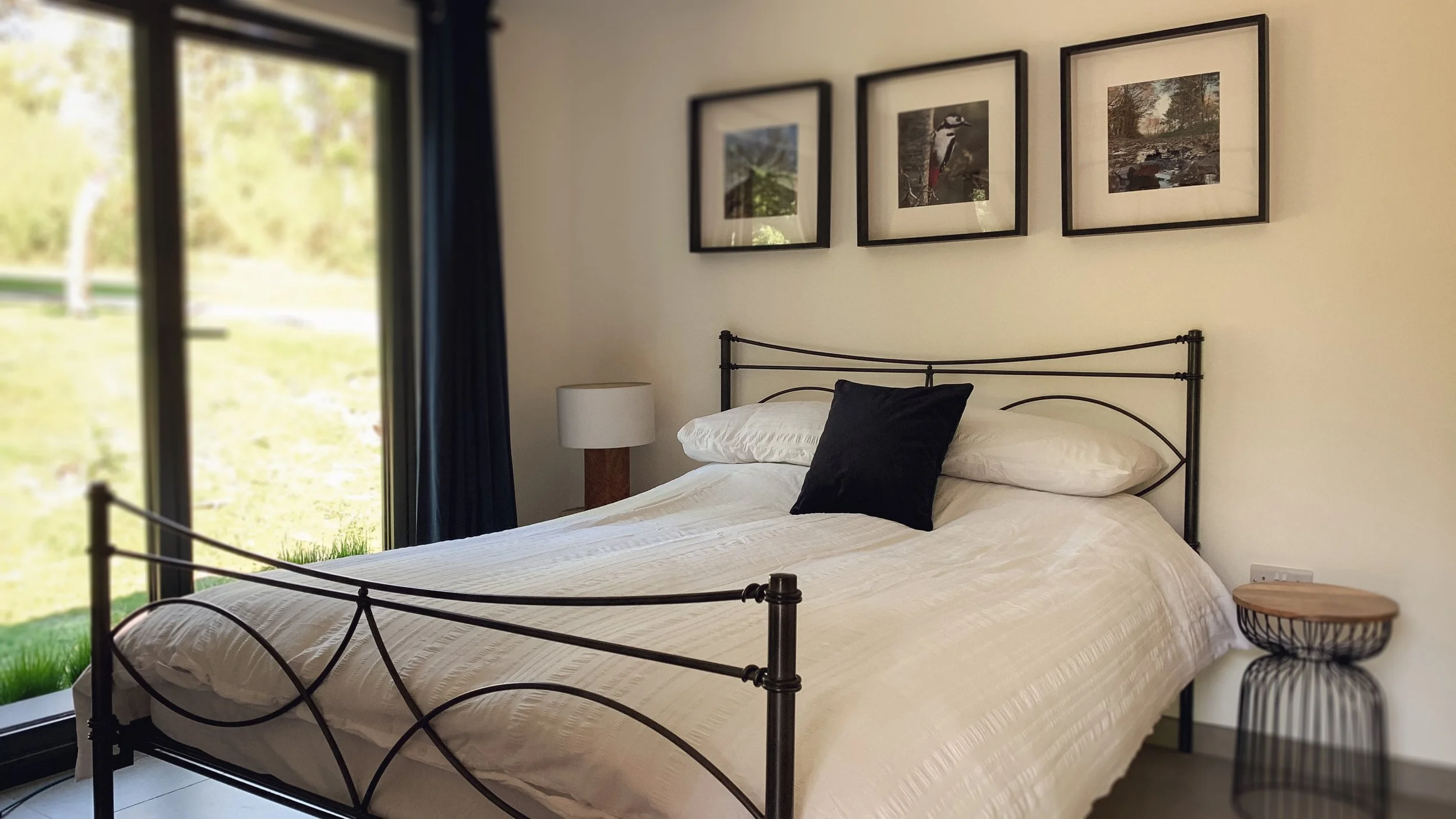 A bedroom with a black metal bed frame, white bedding, black throw pillow, side table with lamp, and framed pictures on a white wall, next to a sliding glass door with dark curtains, overlooking a green outdoor area.