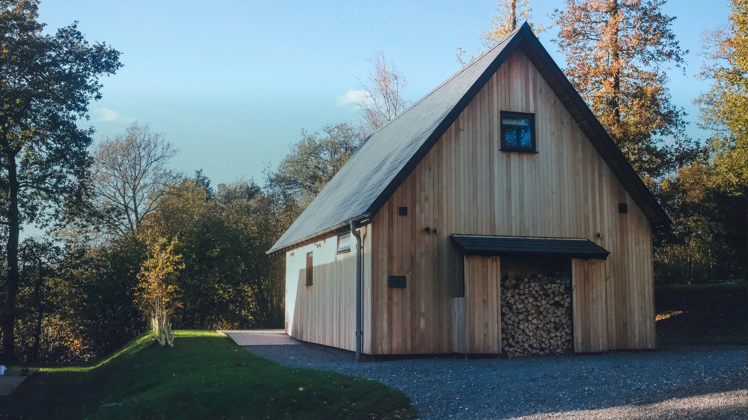 A wooden A-frame house with a steep roof, surrounded by trees with autumn foliage, and a woodpile stored under a small roof extension.