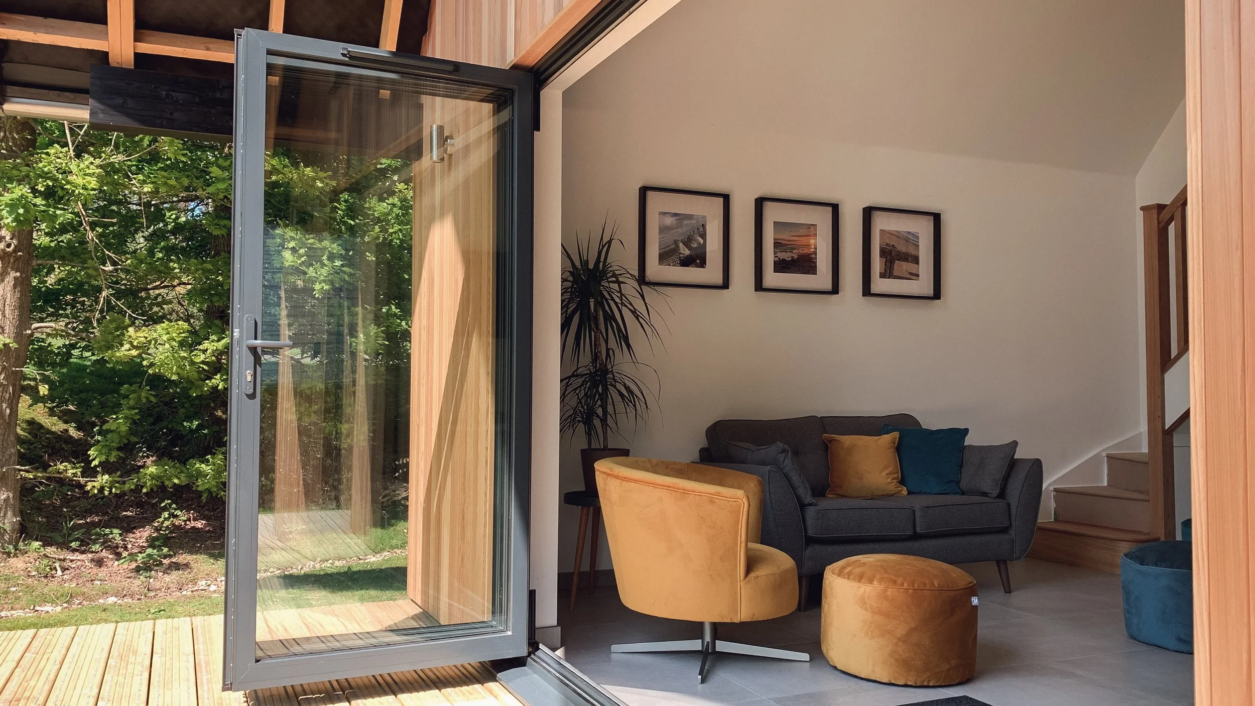 Interior of a modern living room with open glass door leading to a deck and backyard, featuring a gray sofa with yellow and teal pillows, yellow velvet chair and ottoman, framed pictures on the wall, and wooden stairs.