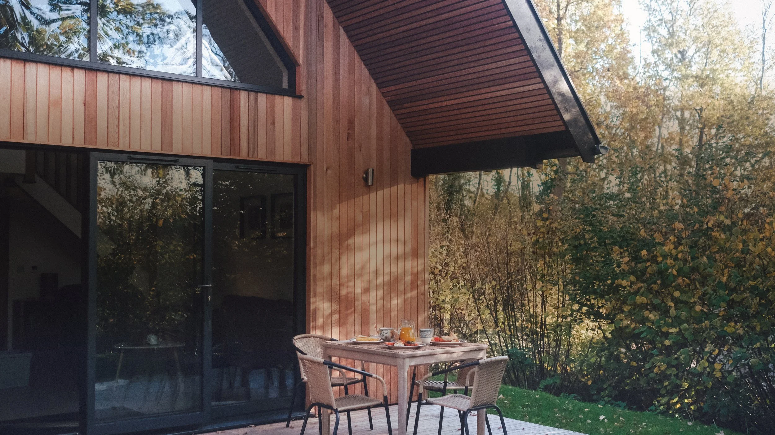 An outdoor patio with a table set for breakfast with bowls, plates, and a jug of orange juice, in front of a modern house with wooden siding and large glass sliding doors, surrounded by trees with fall foliage.