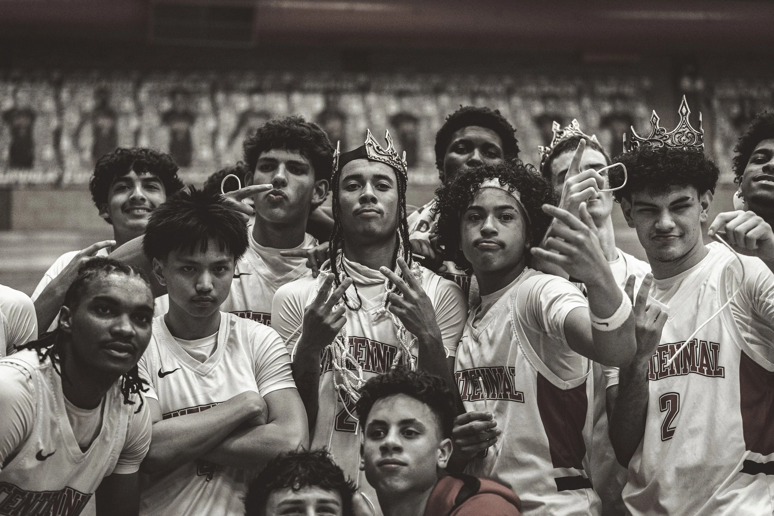 A group of young male athletes wear white sports jerseys, some with crowns, taking a team photo in a stadium. The photo is in black and white.
