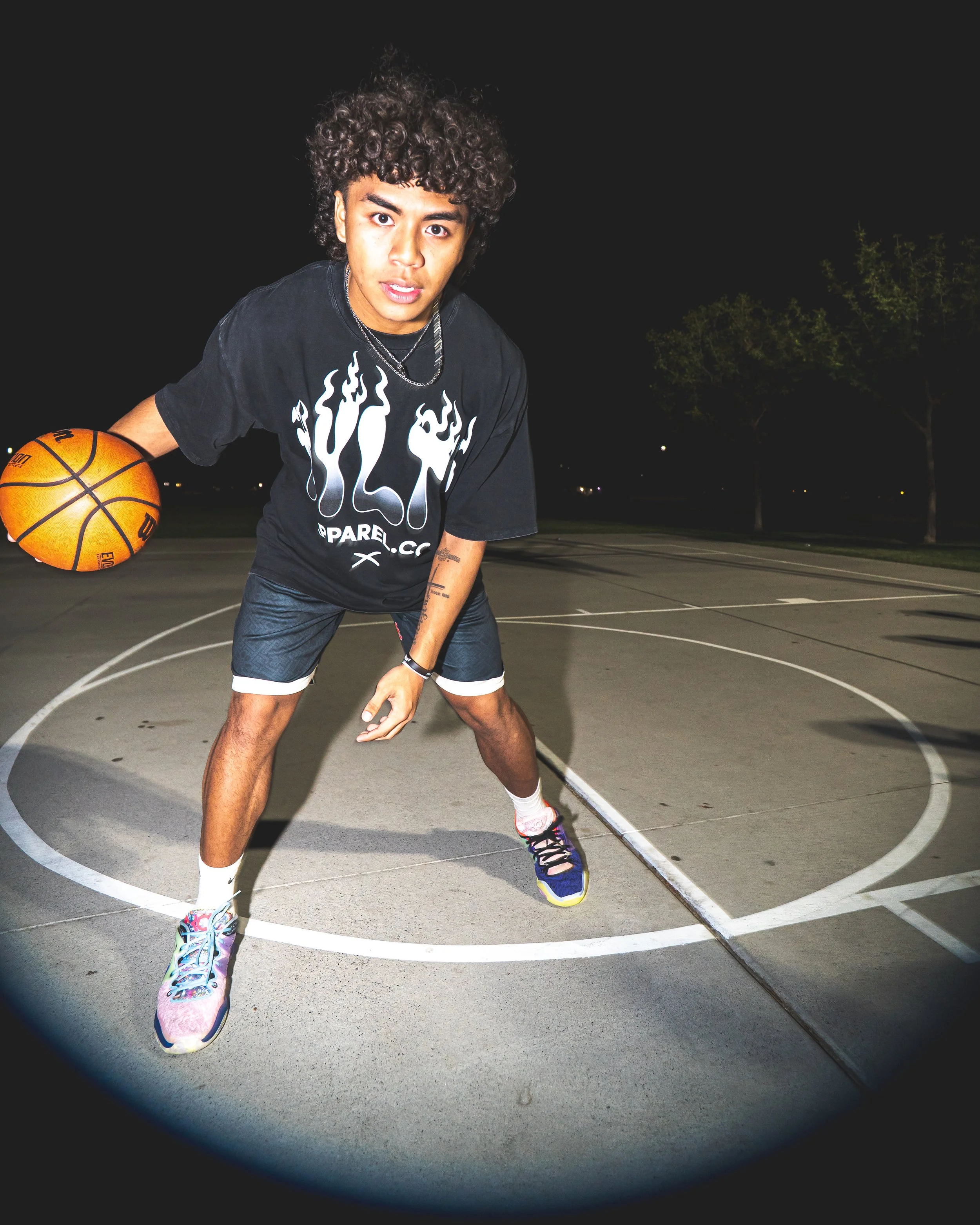 A young man with curly hair is holding a basketball on a basketball court at night. He is wearing a black t-shirt, black shorts, colorful sneakers, and has tattoos on his arm. The court is outdoors with trees and lights in the background.