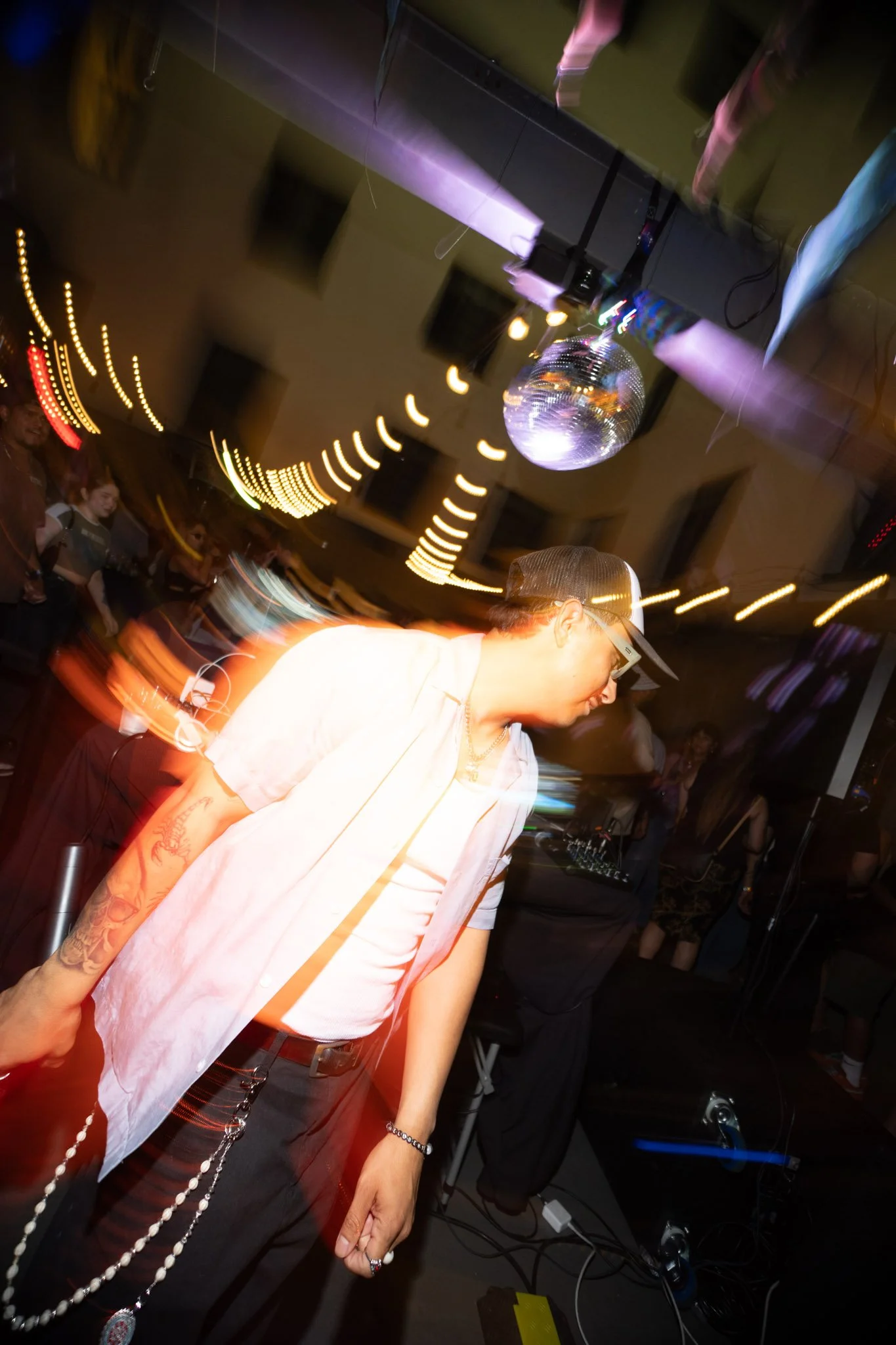 A person with tattoos on their arm, wearing a baseball cap, glasses, a light-colored shirt, black pants, and chains, stands at a dance party with a disco ball hanging from the ceiling, colorful lights, and a crowd of people in the background.