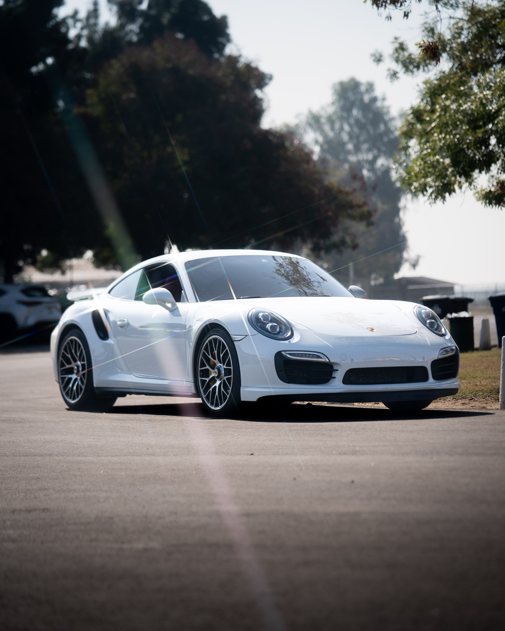 A white sports car parked on a paved area with trees and a cloudy sky in the background.