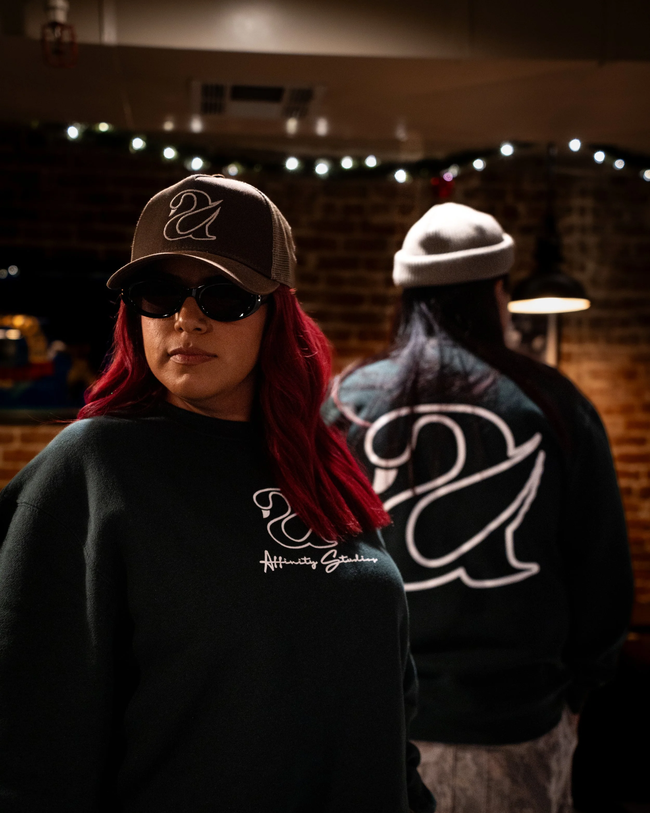 A woman with red hair wearing a gray baseball cap with a swan logo and dark sunglasses. She is wearing a black sweatshirt with white script writing. The background includes another person in gray clothing and a brick wall with Christmas lights.