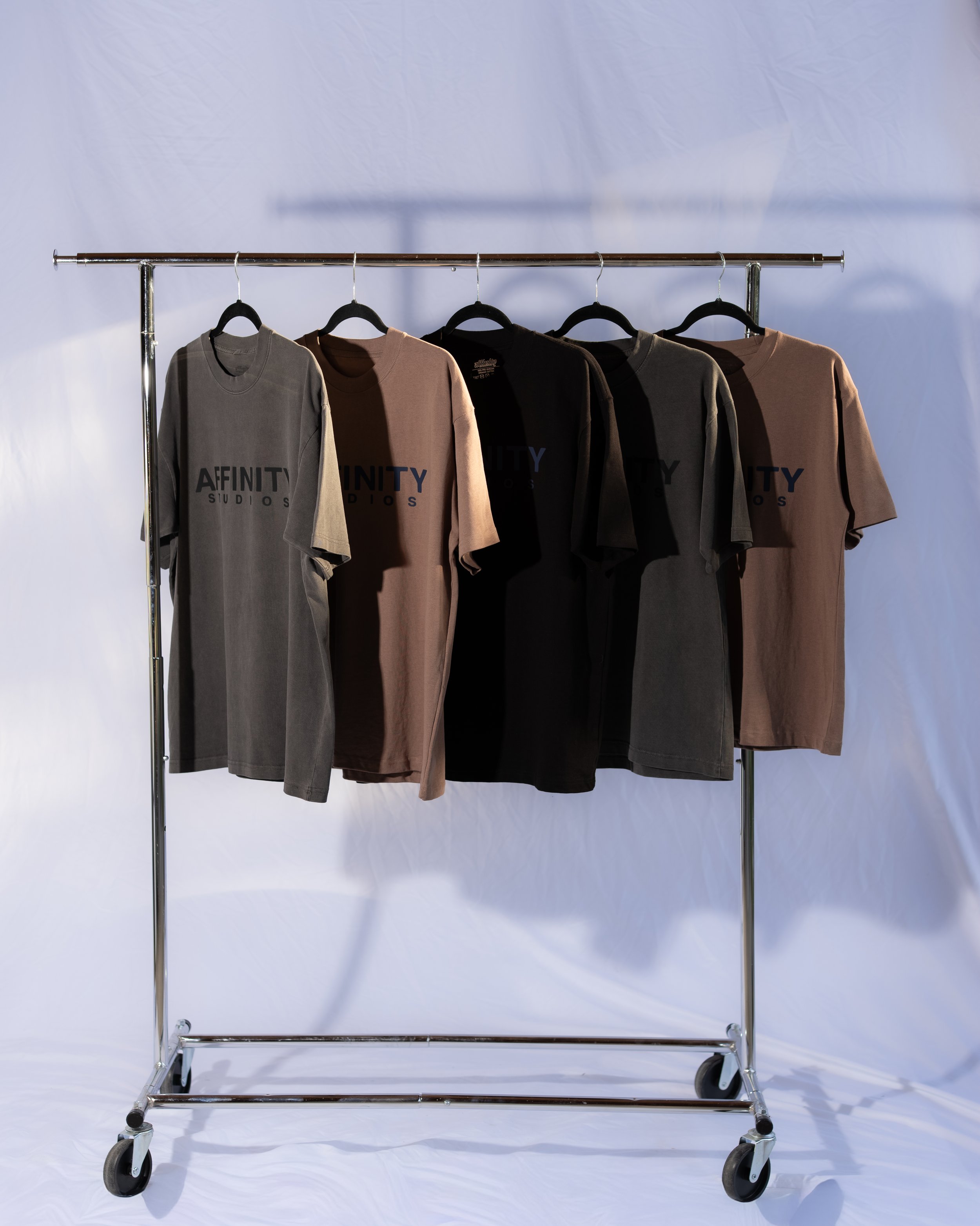 A metal clothing rack on wheels holding five T-shirts hanging on black hangers. The shirts are in beige, brown, black, gray, and beige colors, each with the words 'AFFINITY STUDIOS' printed on the front.