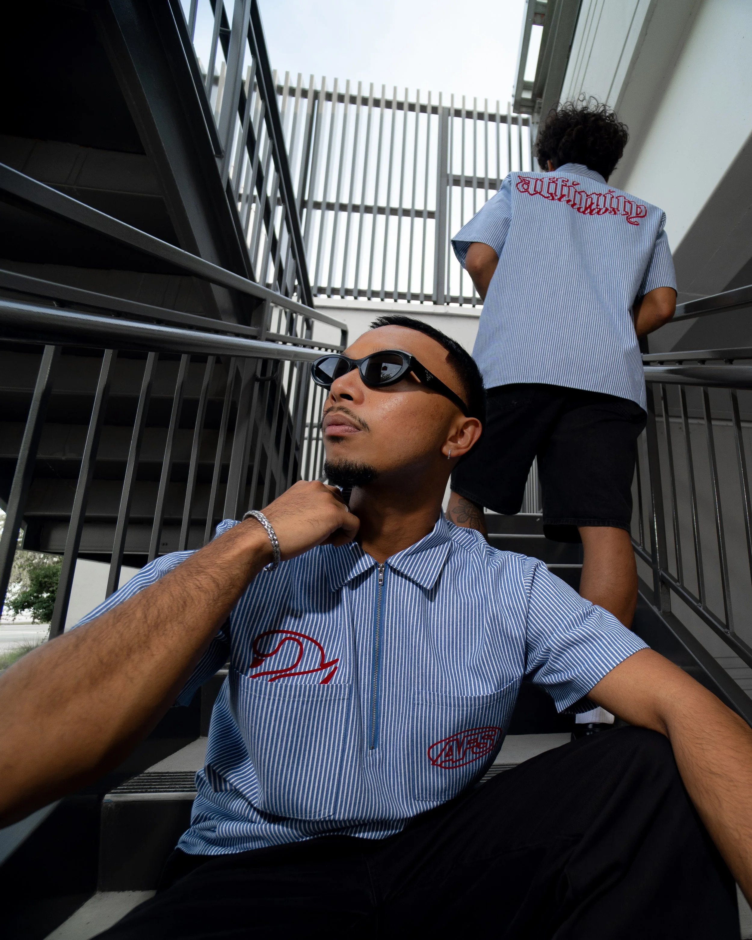 A person wearing sunglasses and a striped shirt sitting on the stairs, with another person standing on the stairs in the background.