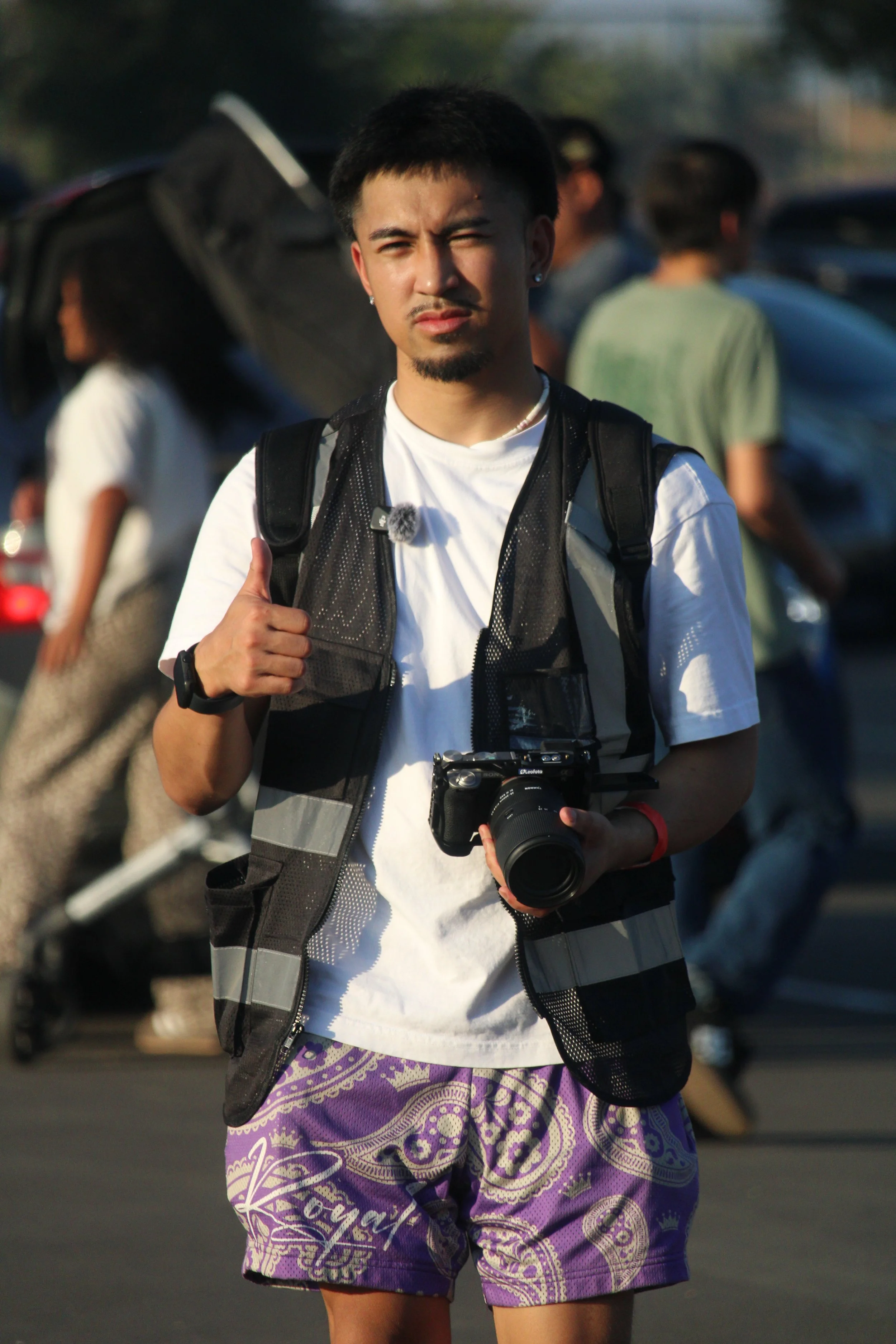 Young man with short black hair, goatee, earrings, wearing a white t-shirt, purple patterned shorts, and a black backpack, holding a camera, with a thumbs-up gesture, outdoors on a sunny day.