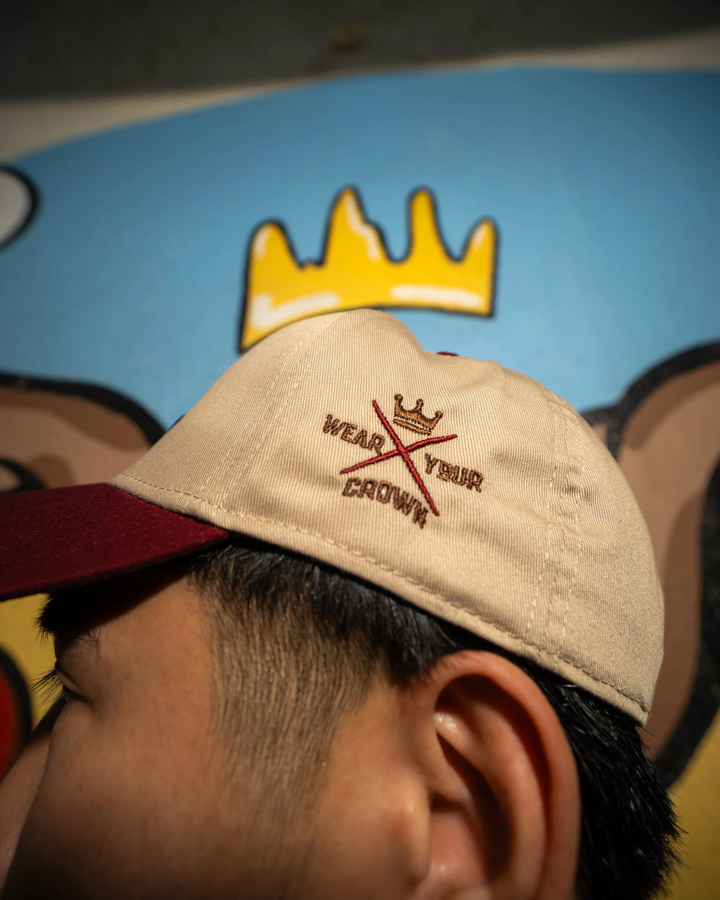 A person wearing a beige baseball cap with a logo that says "We Are Busy Building Future" accompanied by a yellow crown symbol. The person has black hair and is wearing glasses with red frames. The background shows a blue surface with a yellow crown 