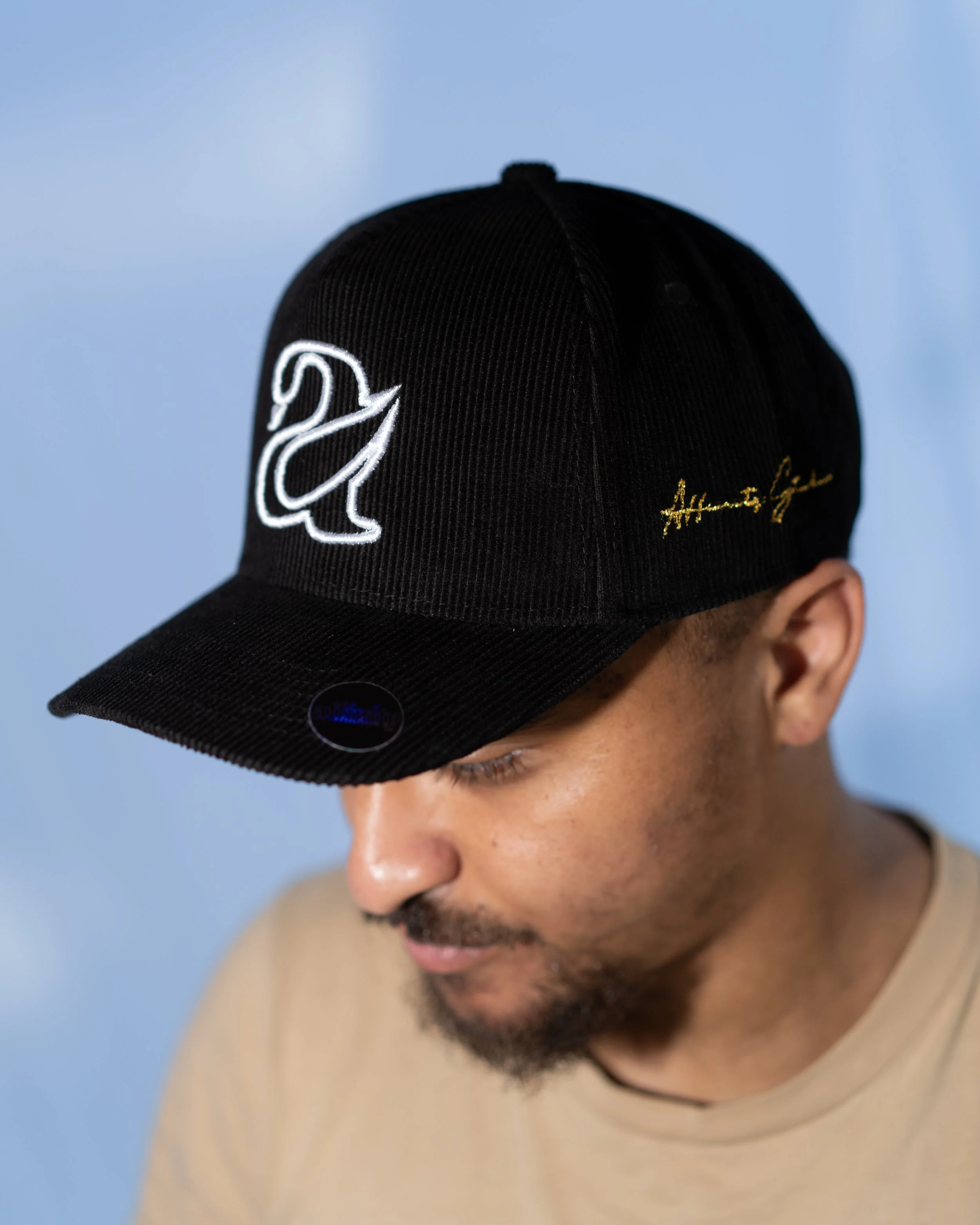 A man wearing a black baseball cap with white and gold embroidery, looking downward, against a blue and beige background.