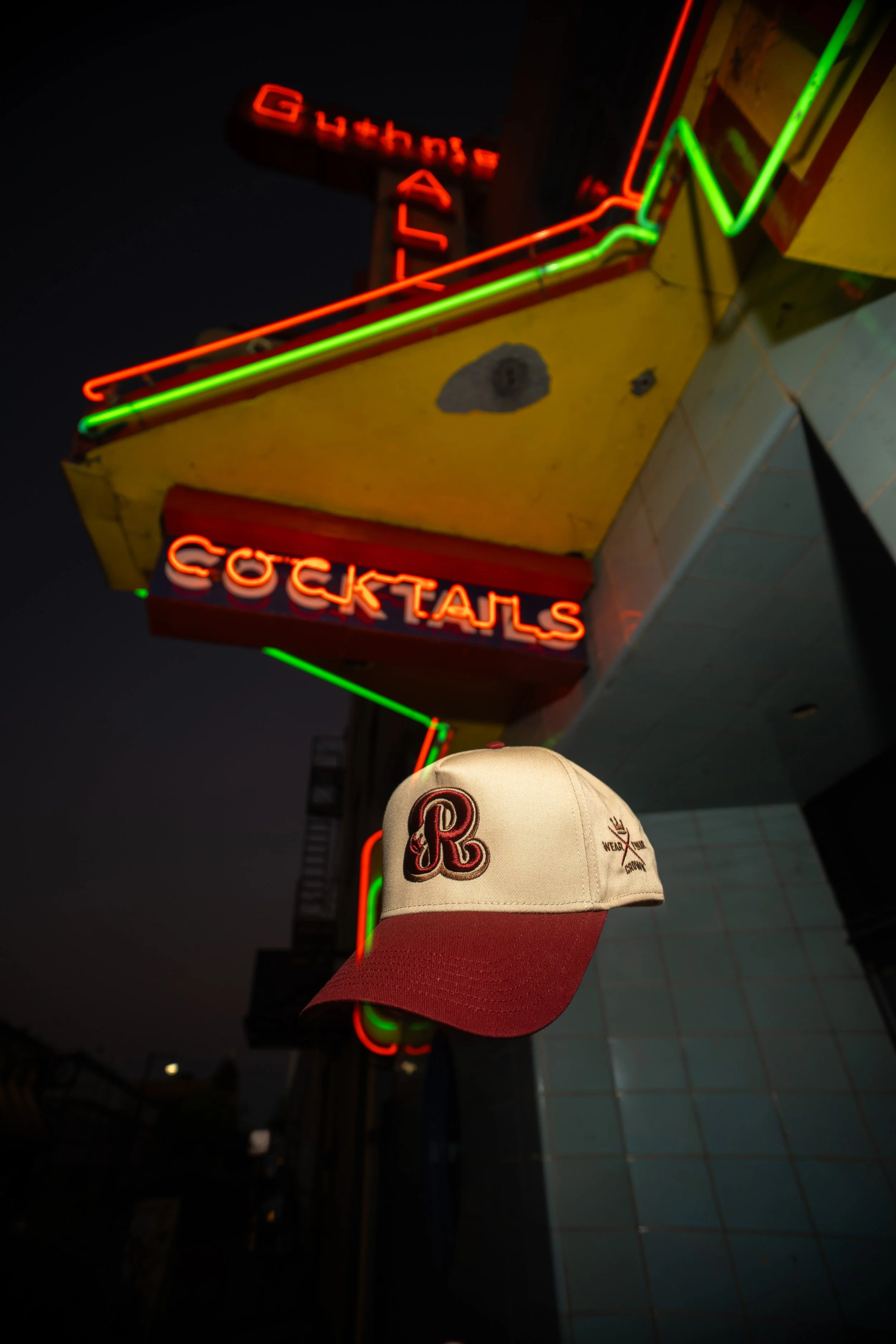 Neon sign with the words 'Coktails' and 'Gitanas' in red and green lights, a white baseball cap with red brim and red embroidered letter 'R' and a small logo, hung on the wall in front of a bathroom mirror at night.