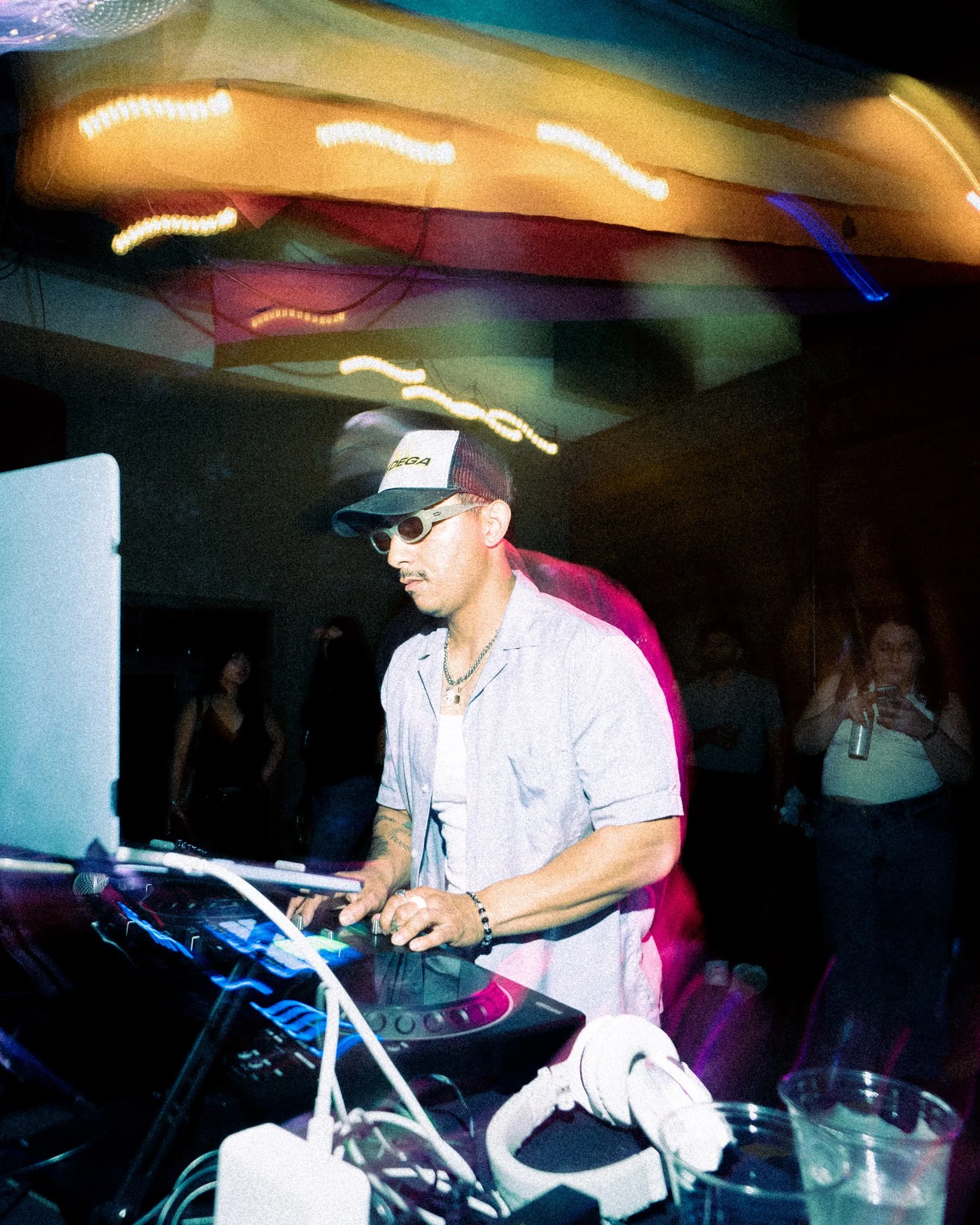 A man wearing a baseball cap, sunglasses, and a light-colored blazer is DJing at a party or nightclub, standing behind DJ equipment with colorful lights reflecting above.