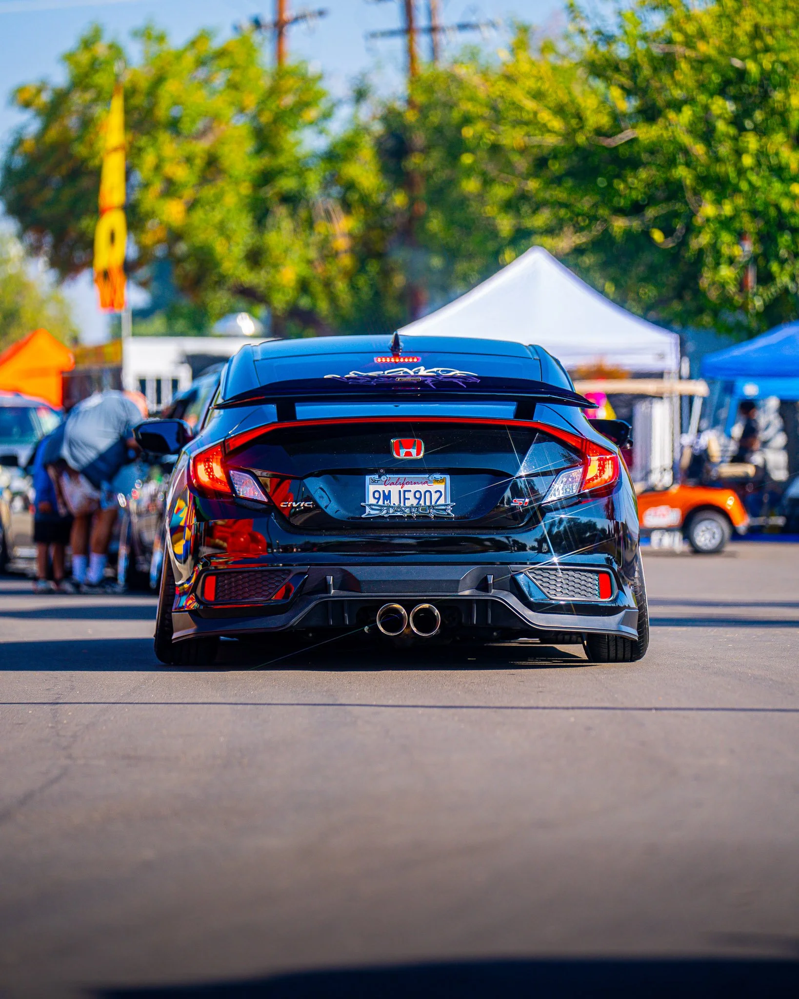 Black Honda Civic car with dual exhaust pipes and California license plate parked outdoors at an event or market, with tents and trees in the background.