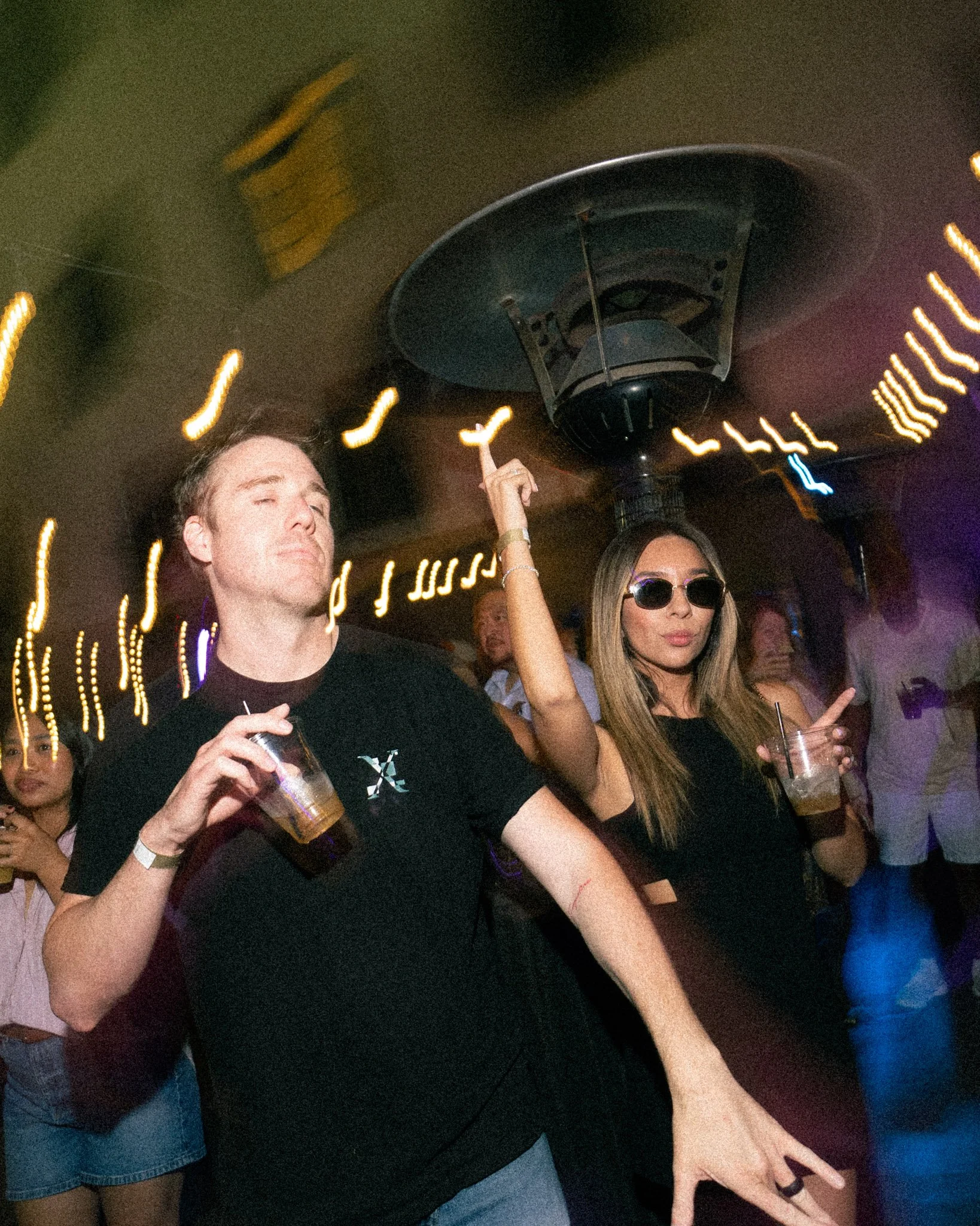 People at a nightclub or party holding drinks, with colorful lights and dancing in the background.