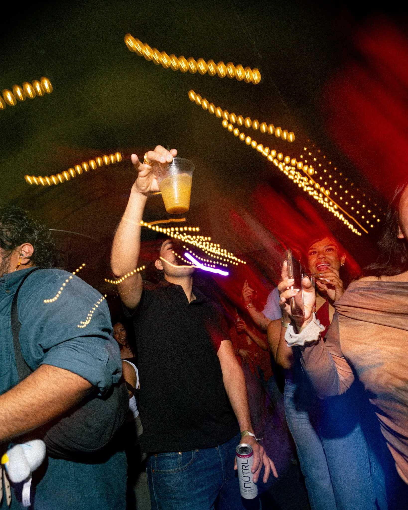 People at a party with festive string lights overhead. One person is holding a drink in a clear cup, and some individuals are holding cans and glasses. The scene appears lively and cheerful.
