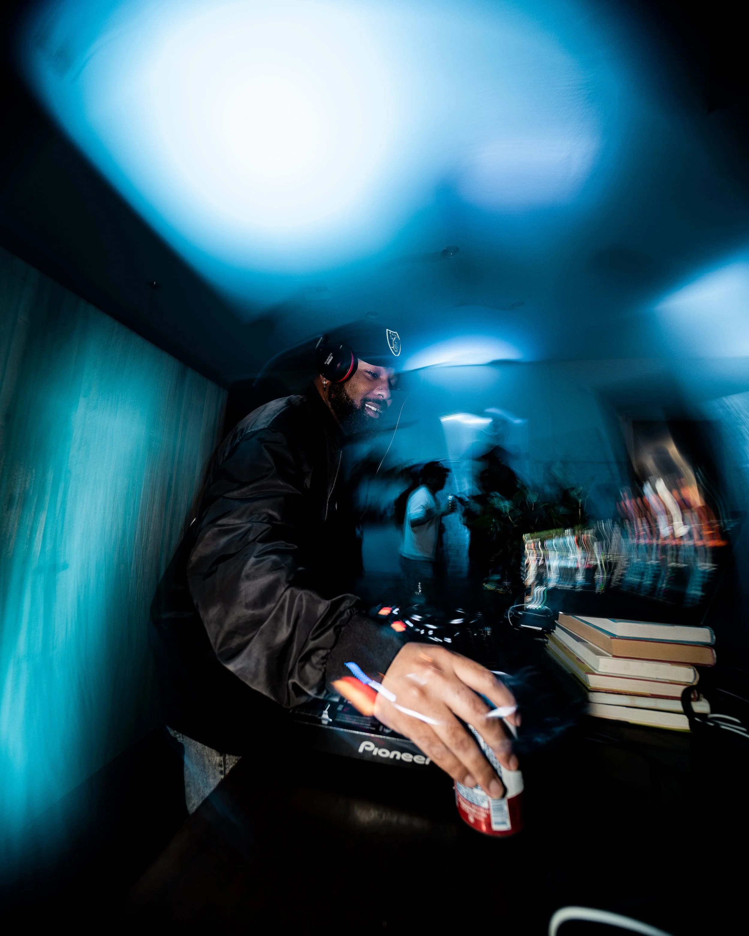 A man smiling while DJing with headphones on, using a Pioneer DJ controller, in a dimly-lit room with blue lighting and books on a table.