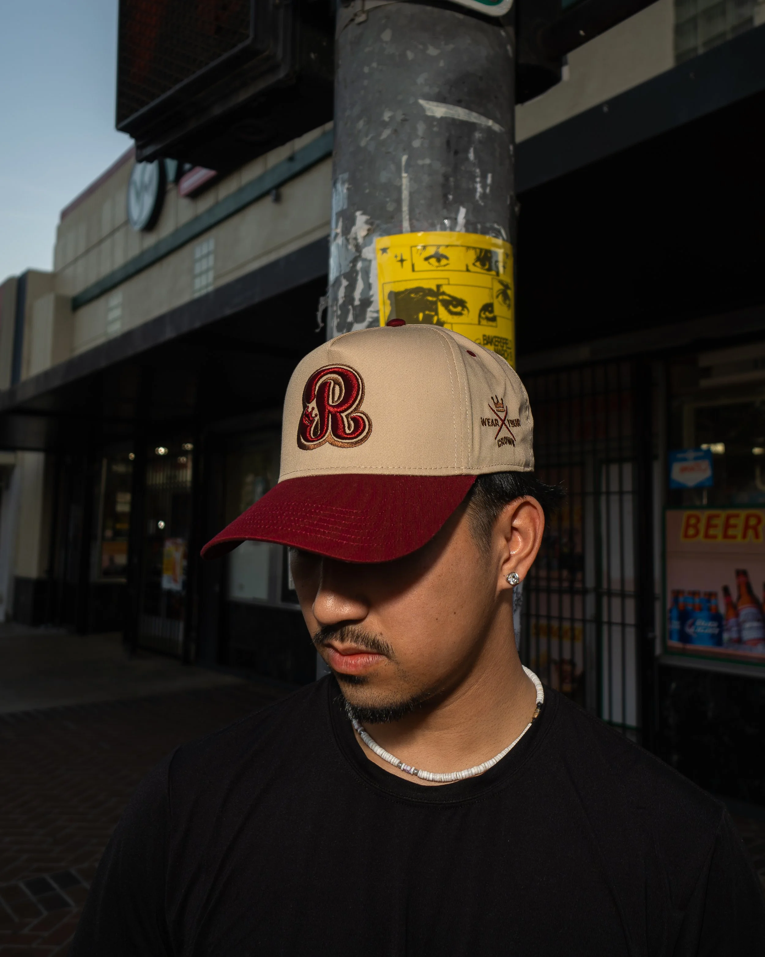 A person wearing a beige cap with a red brim and embroidered 'R' and a black t-shirt, standing outdoors near a pole with a yellow informational sticker, with a building and a sign in the background.
