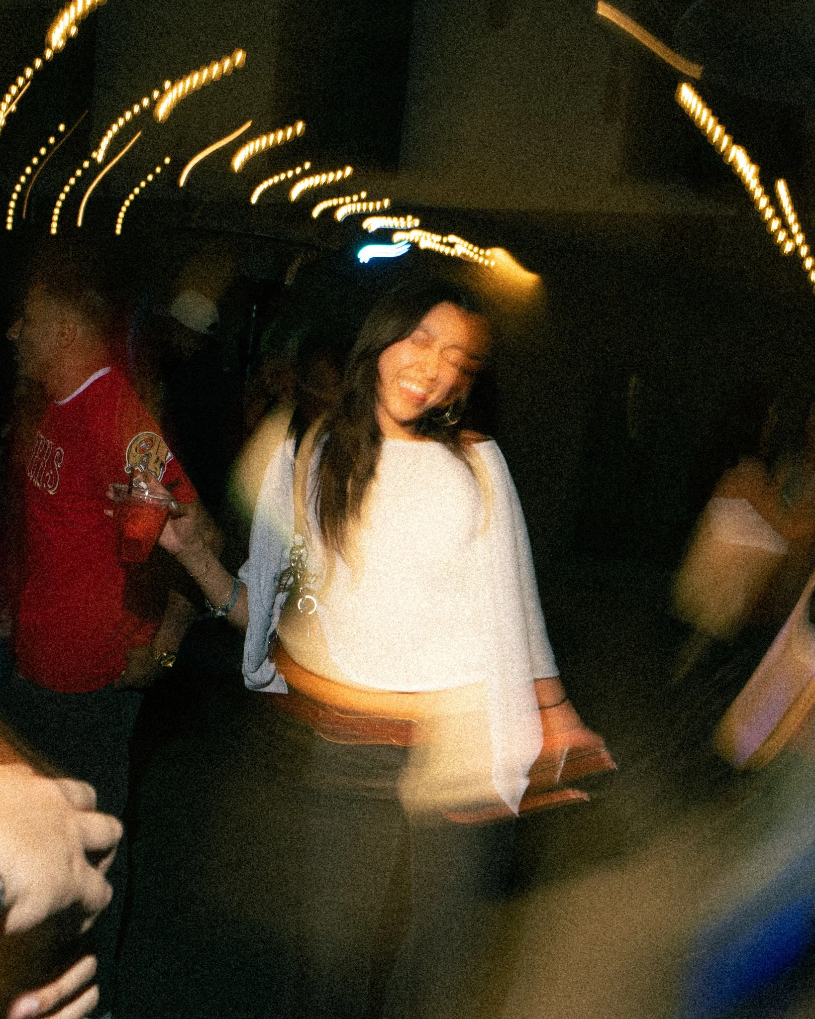 Smiling woman with long dark hair wearing white top, in a dimly lit space with string lights overhead, near a person in a red shirt.