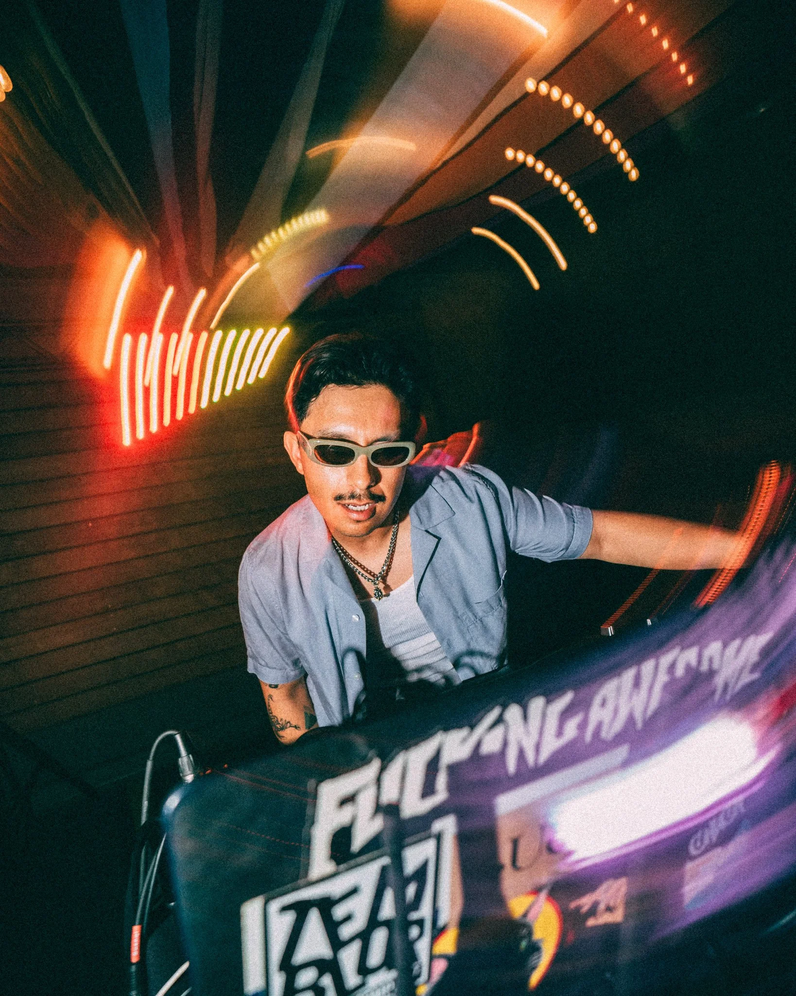 A man wearing sunglasses and a chain necklace is DJing at a nightclub, with colorful neon lights and a digital sign in the background.