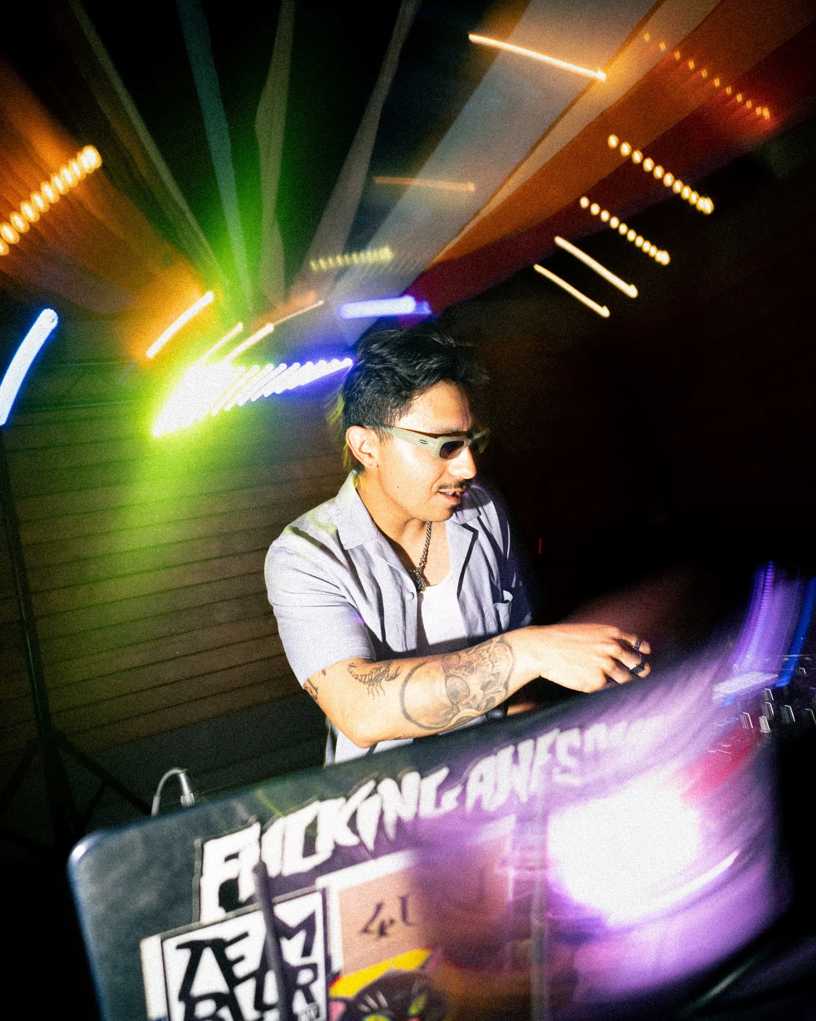 A young man with tattoos on his arm wearing sunglasses and a gray jacket, standing in front of colorful neon lights at night.
