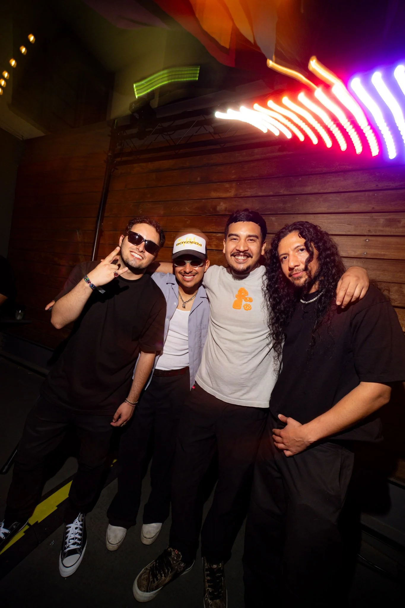 Four men posing for a photo at a party or event, standing in front of a wooden wall with colorful, bright light trails above them. One man is wearing sunglasses and making a hand gesture, another with a cap labeled 'BODEGA.' They are smiling and appe