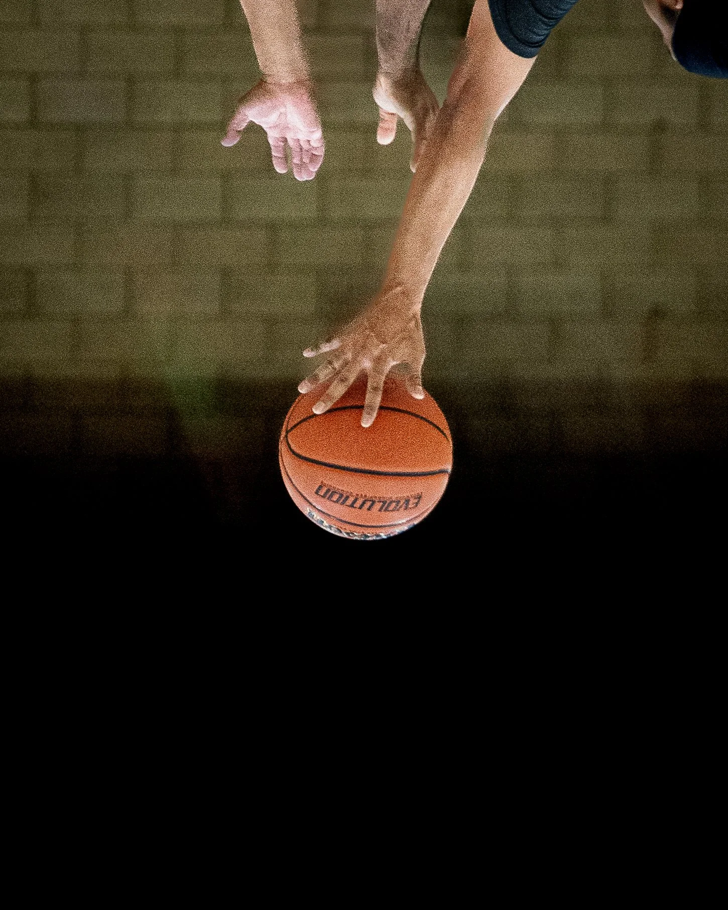 A person reaching for a basketball on a dark court with their right hand, with the person's left hand hanging near the floor.