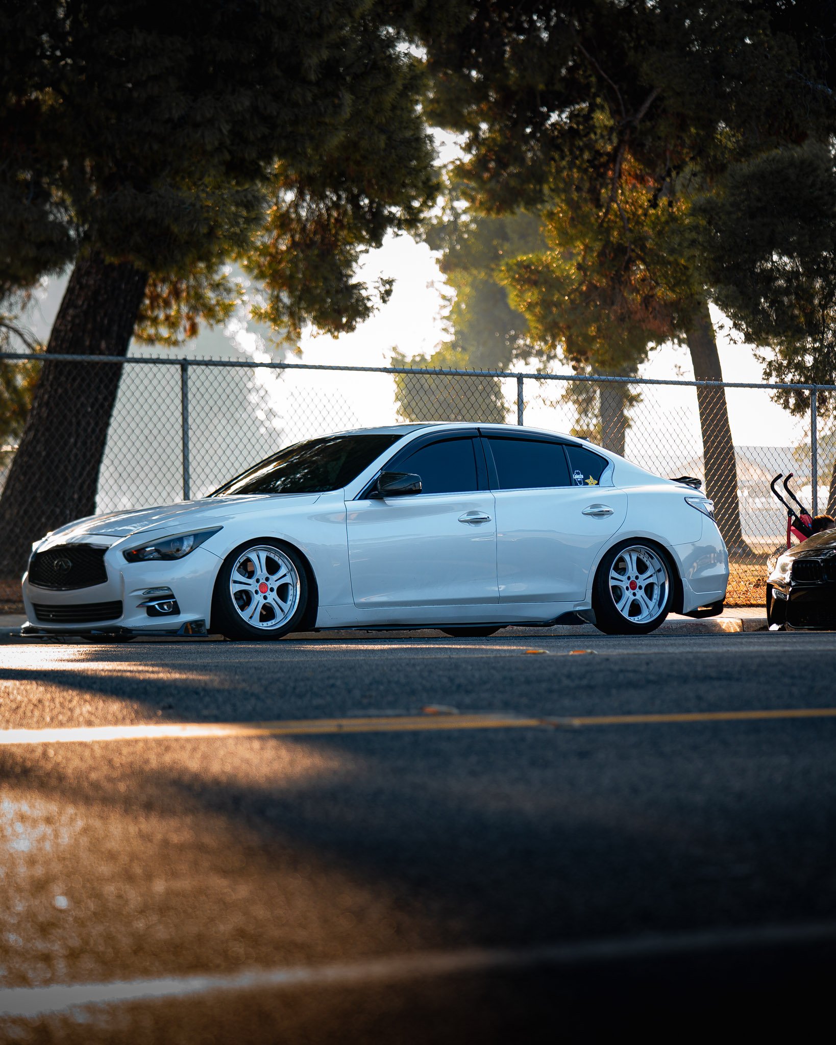 Side view of a white car parked near a chain-link fence, with large trees in the background during sunset or sunrise.
