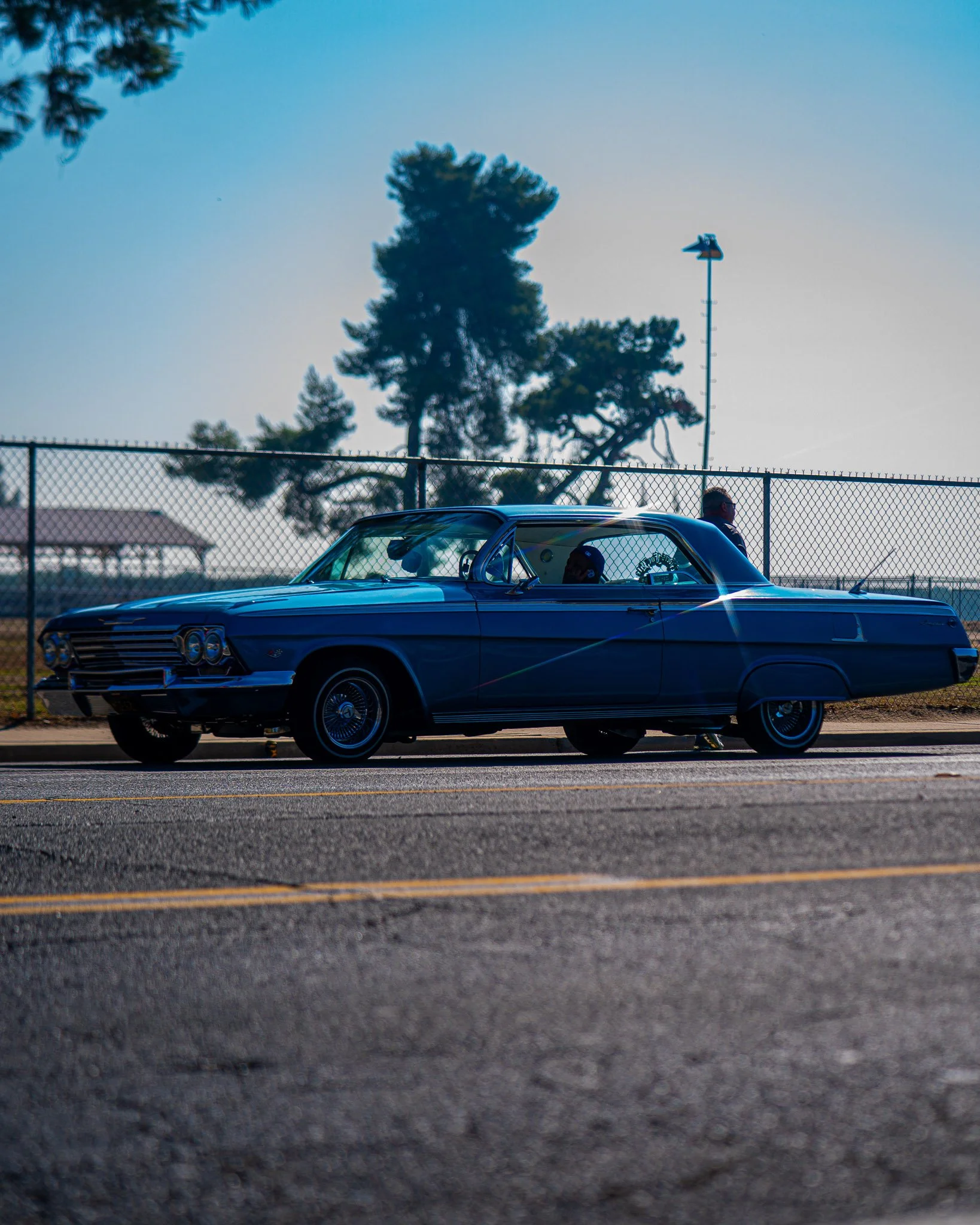 A classic blue car driving on the road near a chain-link fence with a person sitting inside, possibly a driver. In the background, there are trees, a lamppost, and a small building or gazebo under a clear sky.
