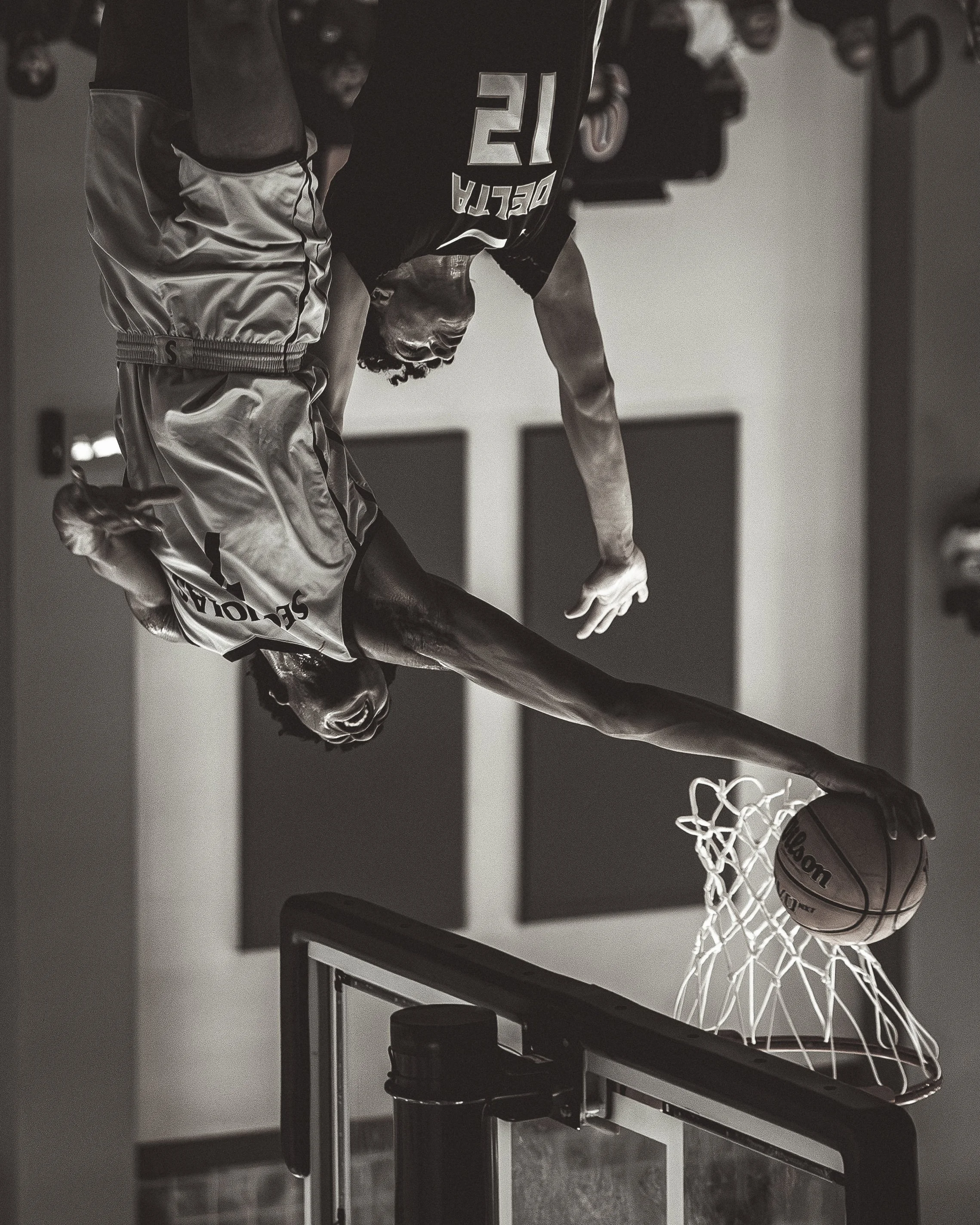A person playing basketball indoors, captured in a black and white photo, with a basketball going into the hoop. The person is reaching towards the hoop and wearing shorts and a sleeveless top.