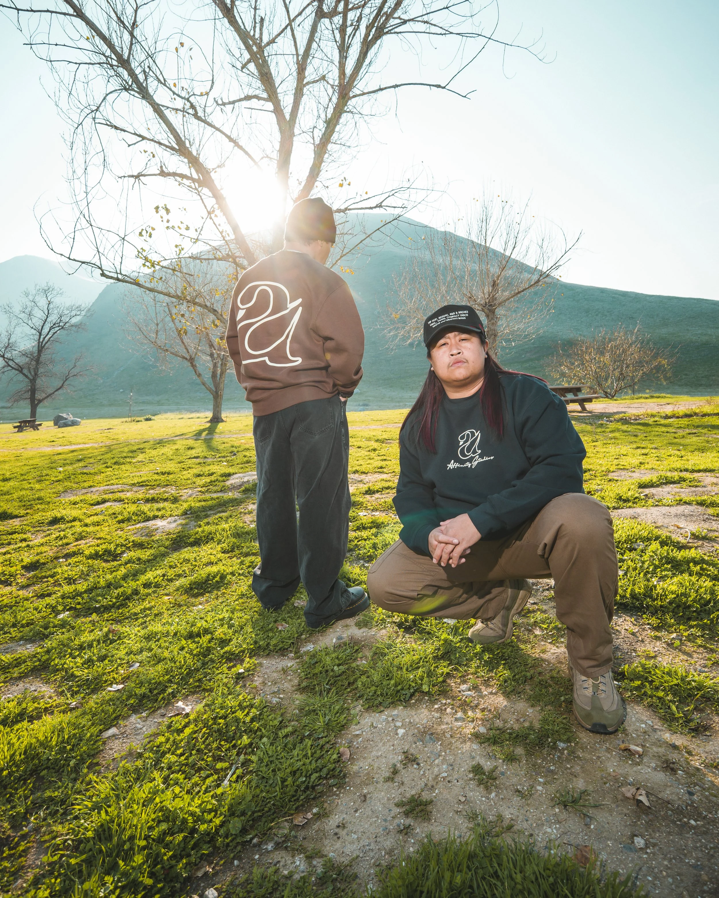 Two people outdoors in a park during daytime, one standing with their back to the camera and the other kneeling in front. The background includes leafless trees, a grassy area, picnic tables, and mountain hills. The sun is shining brightly, creating 