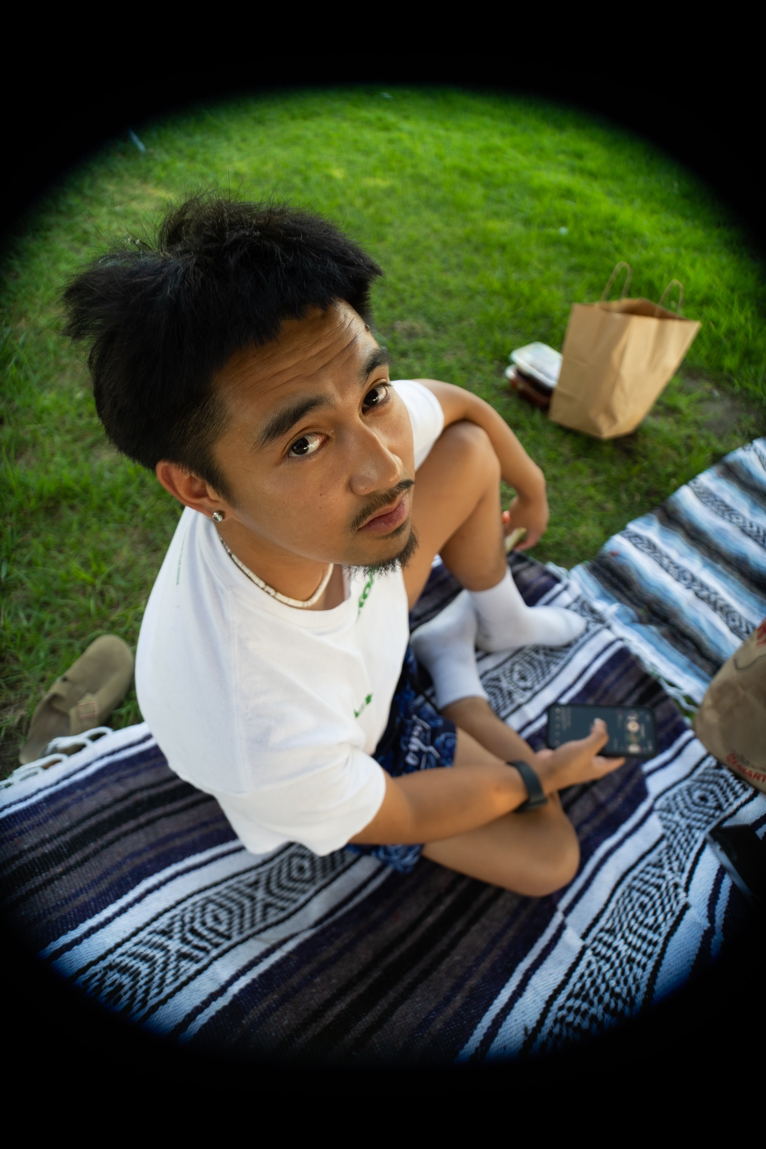 A young man with dark hair and facial hair, sitting on a blanket on the grass, looking up at the camera. He is holding a smartphone in his right hand. In the background, there is a paper bag and some personal items on the grass.