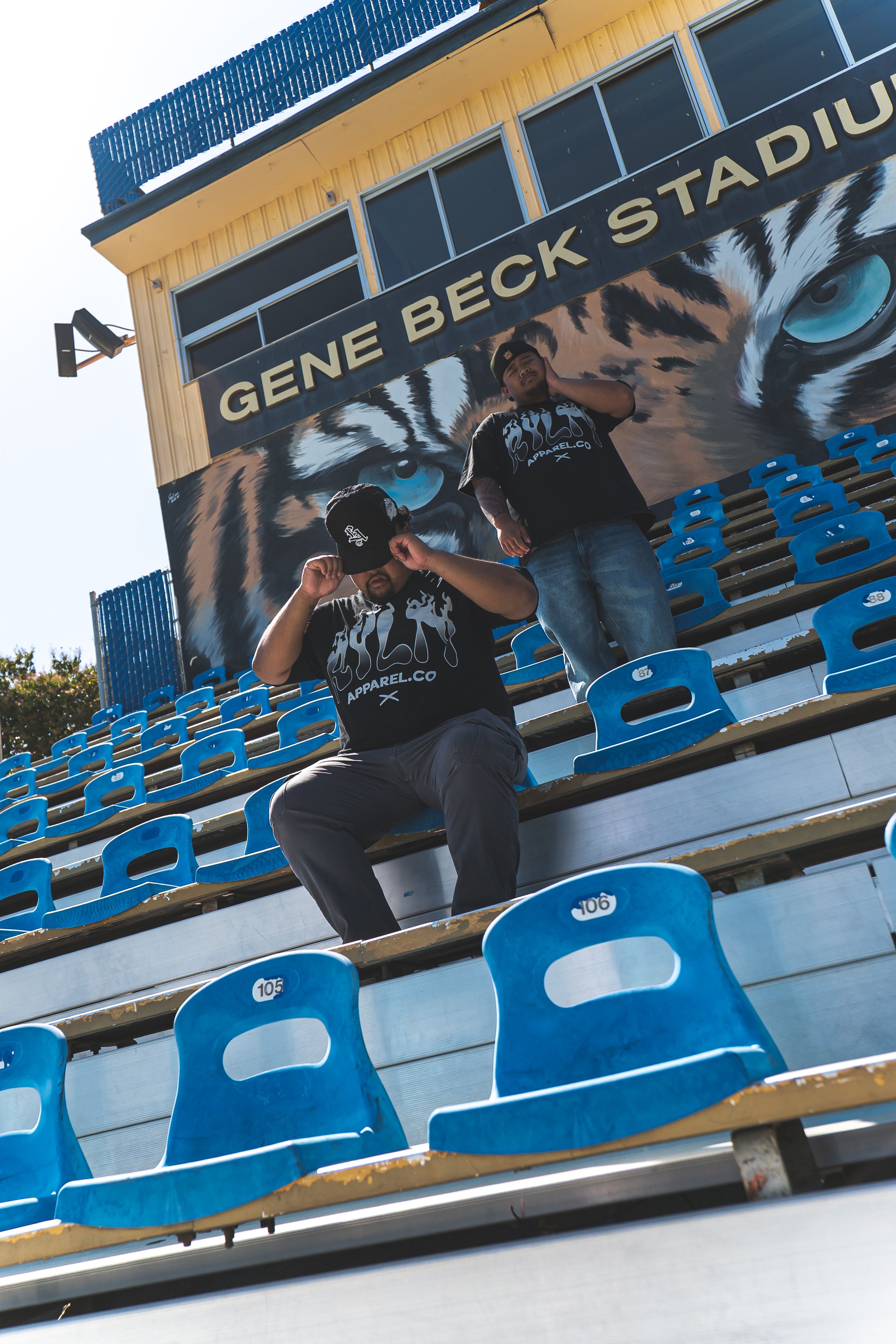 Two young men standing on stadium bleachers in front of a sign that says "Gene Beck Stadium" with a large painted eyes of a tiger below the sign. The bleachers are mostly empty with some blue seats numbered.
